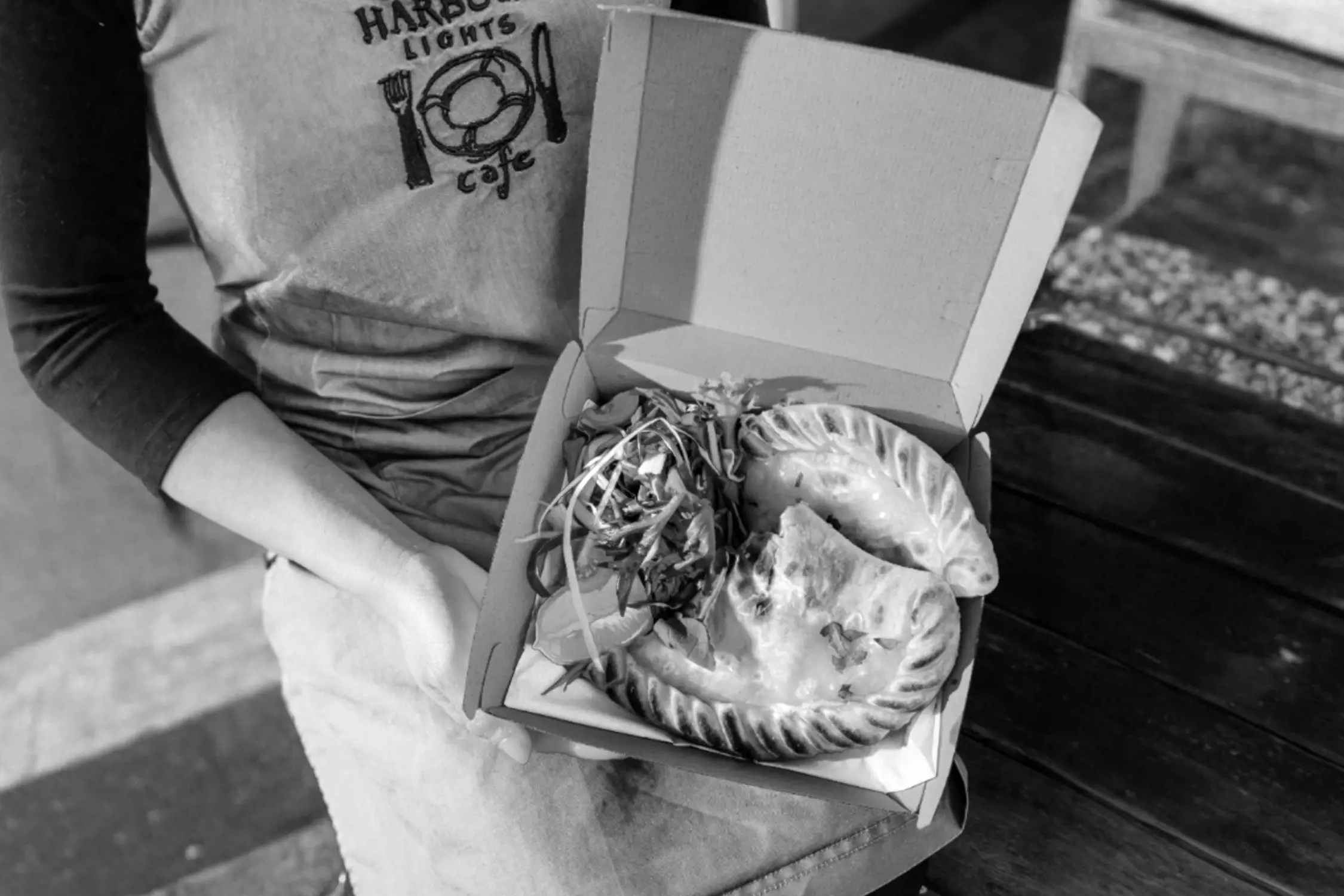 Black and white close-up photo of a person holding a takeout box containing two pasties and a side of salad, seated on a wooden bench.