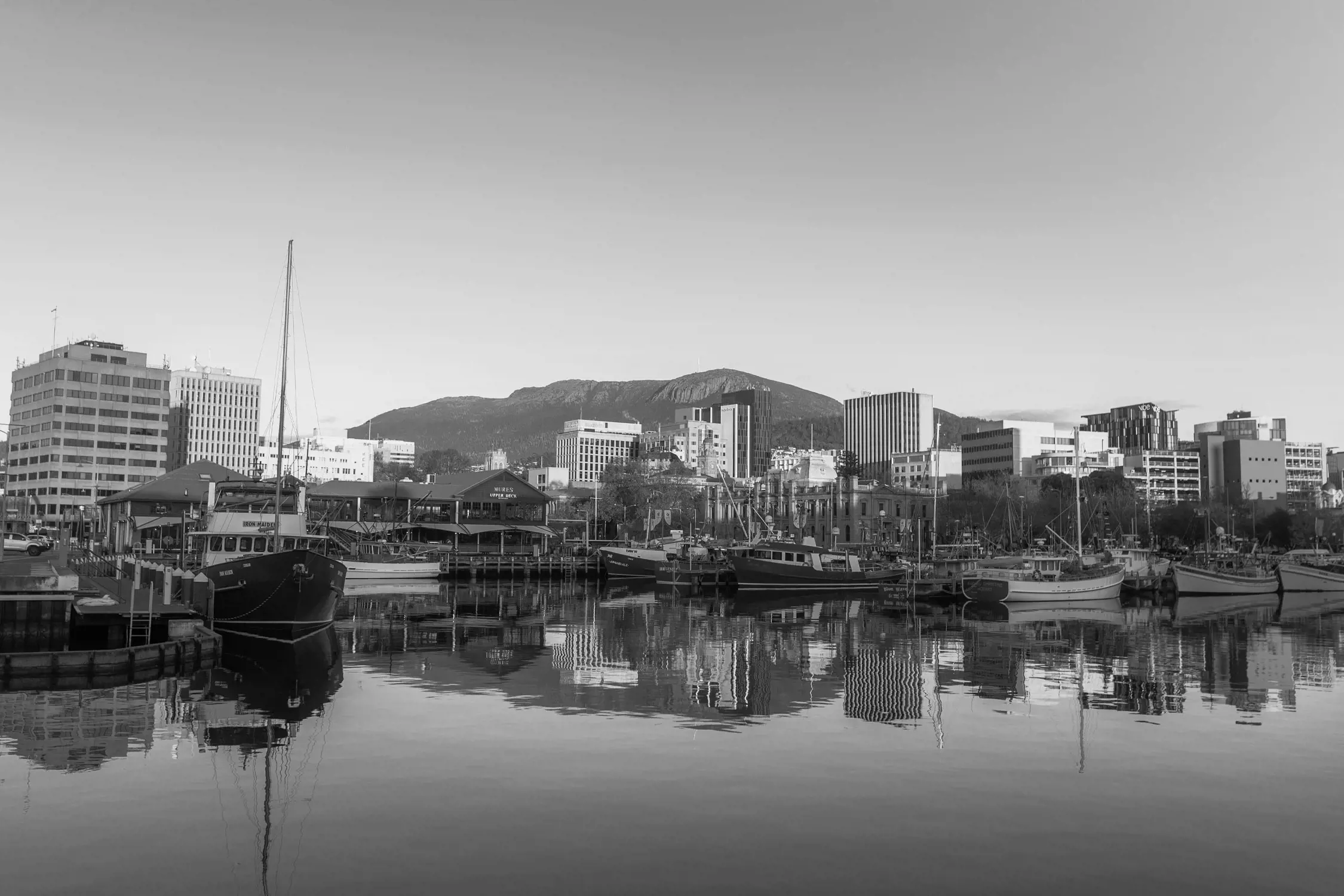 Black and white photo of a harbor scene with boats docked, buildings in the background, and a mountain range behind the city, with calm water reflecting the scene.