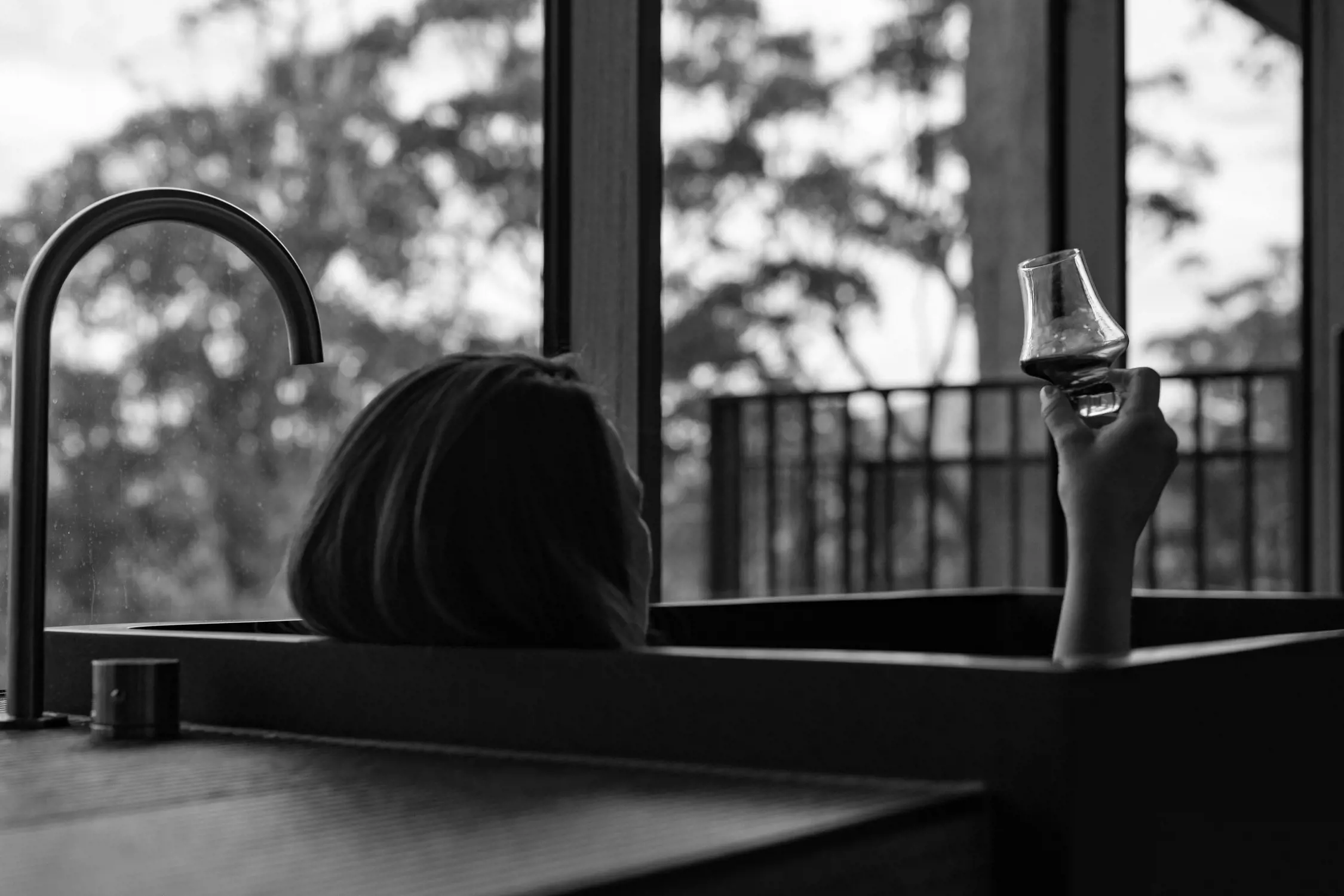 Black and white photo of a person relaxing in a bathtub, holding up a small glass of whiskey or liquor, with a large window and trees visible in the background.