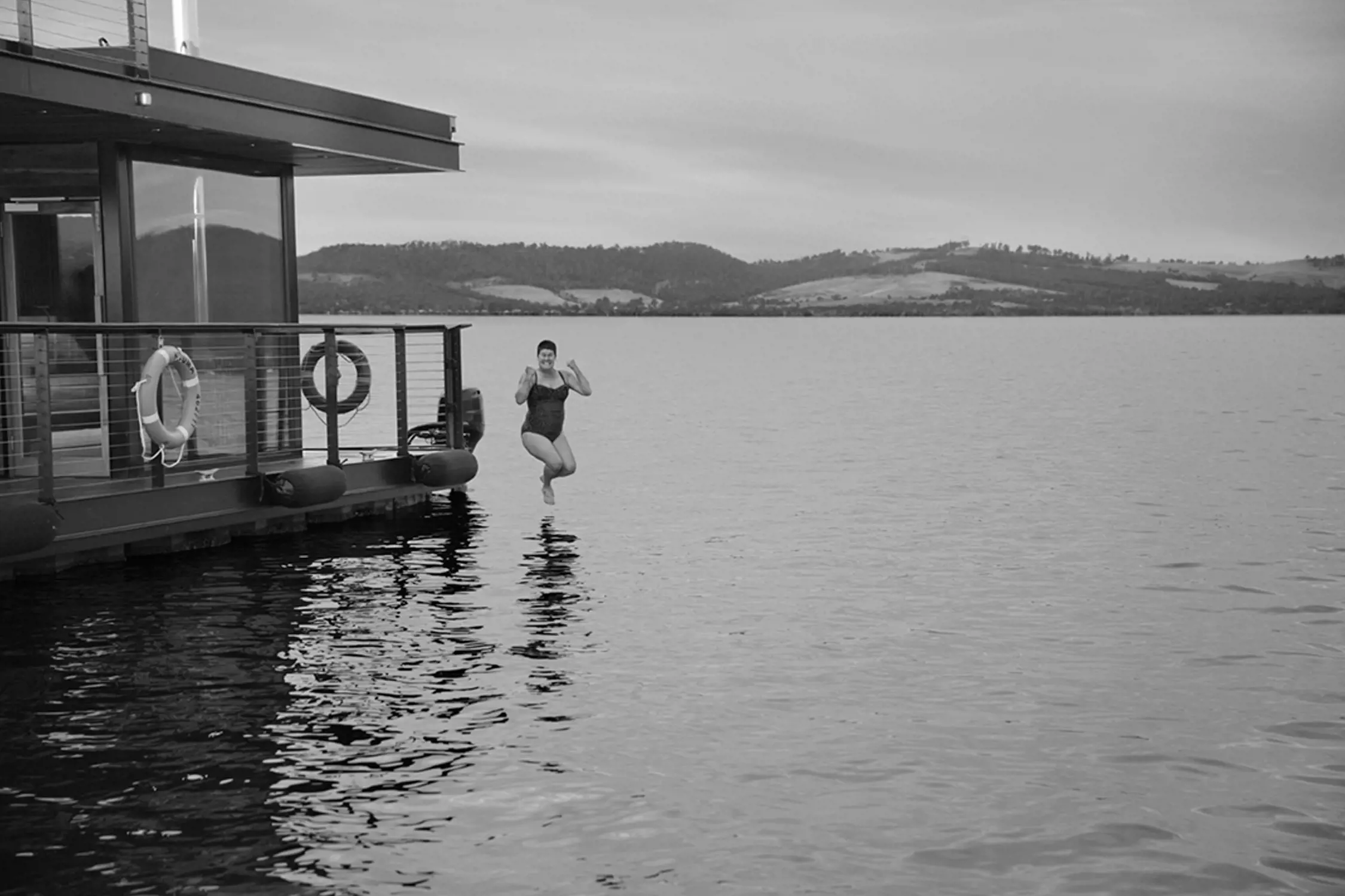 Black and white photo of a person mid-air, jumping off a modern floating dock into a calm lake with hills visible in the background.