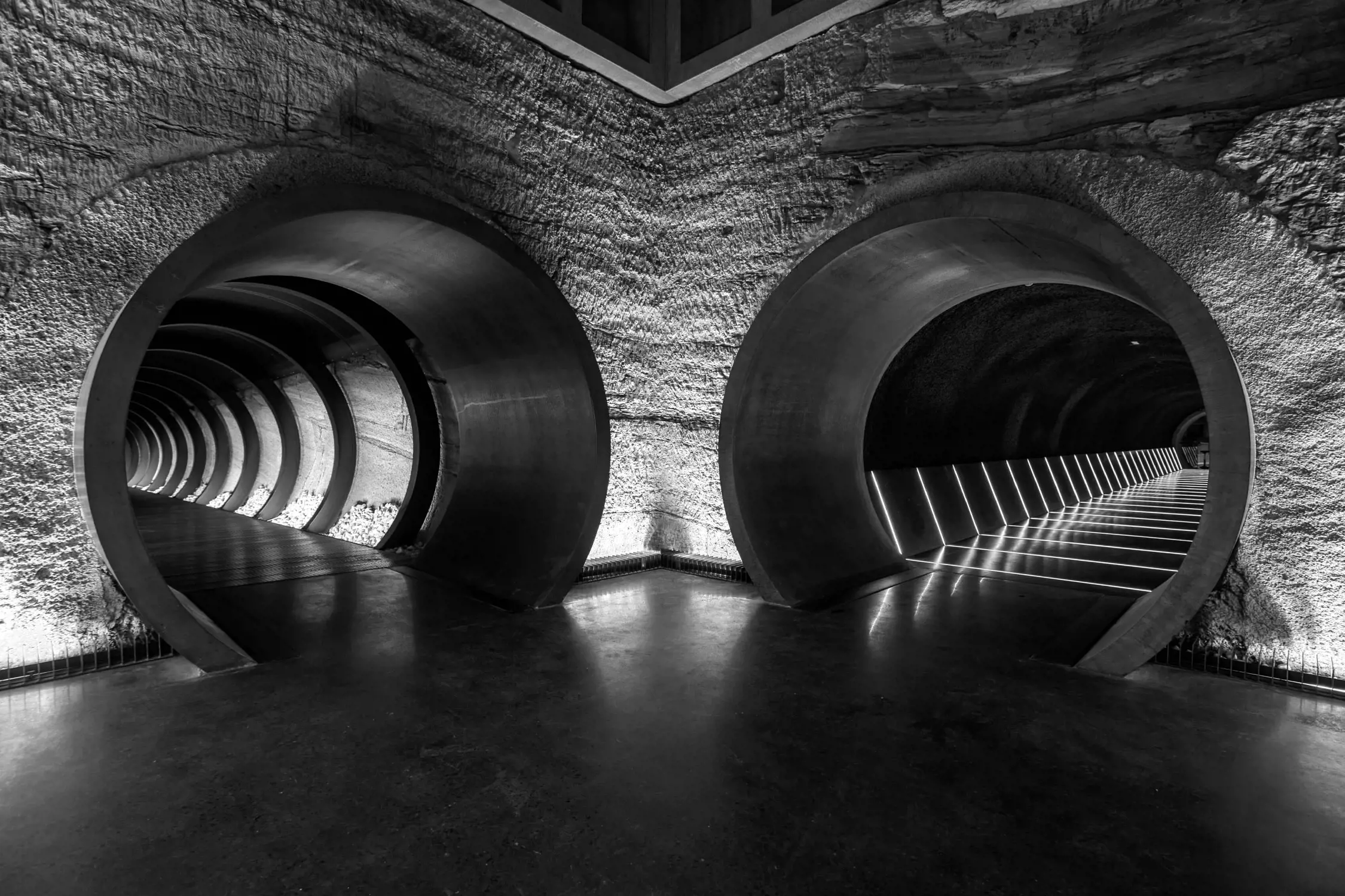 Black and white photo of a symmetrical concrete structure with two large circular tunnels; the tunnel on the left is lit with aligned lights creating a spiraling effect, while the tunnel on the right has linear lights illuminating the pathway.