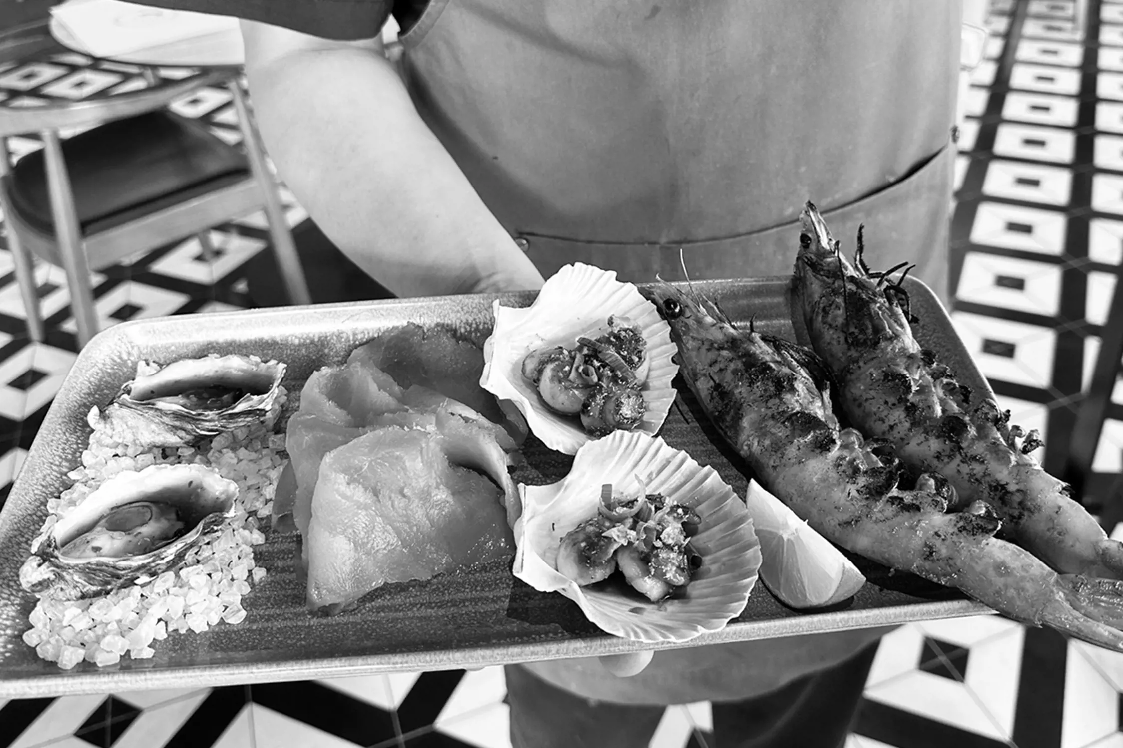 Black and white photo of a tray of assorted fresh seafood including oysters, scallops, prawns, and sashimi held by a person wearing an apron