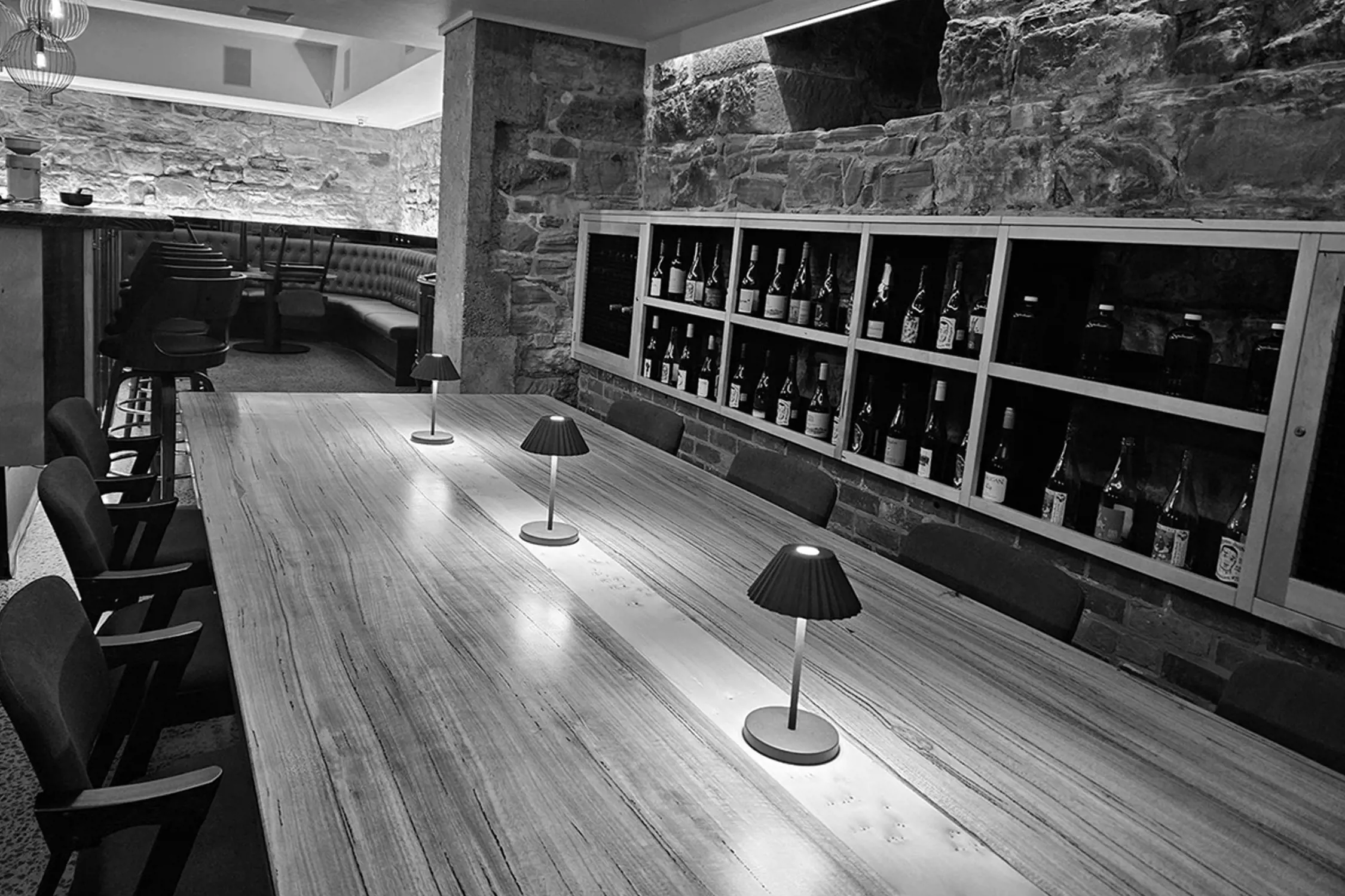 Interior image of a modern tasting room or bar featuring a long wooden table with small lamps, surrounded by chairs, and a wall-mounted wine rack filled with bottles.
