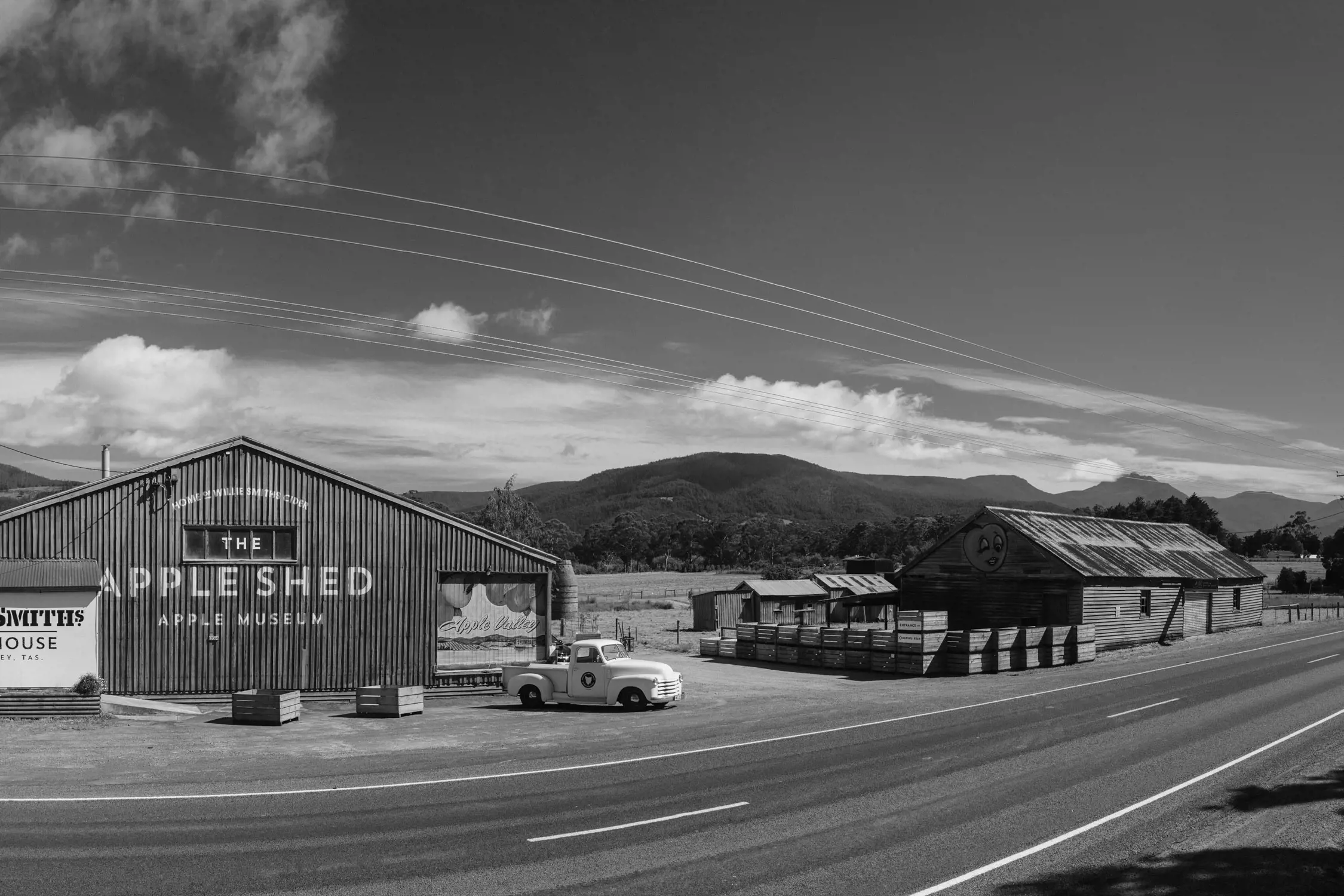 Rural scene showing an old-style wooden building labeled "The Apple Shed Apple Museum" and "Willie Smith's Cider House" with a vintage truck parked outside, set against mountainous countryside.