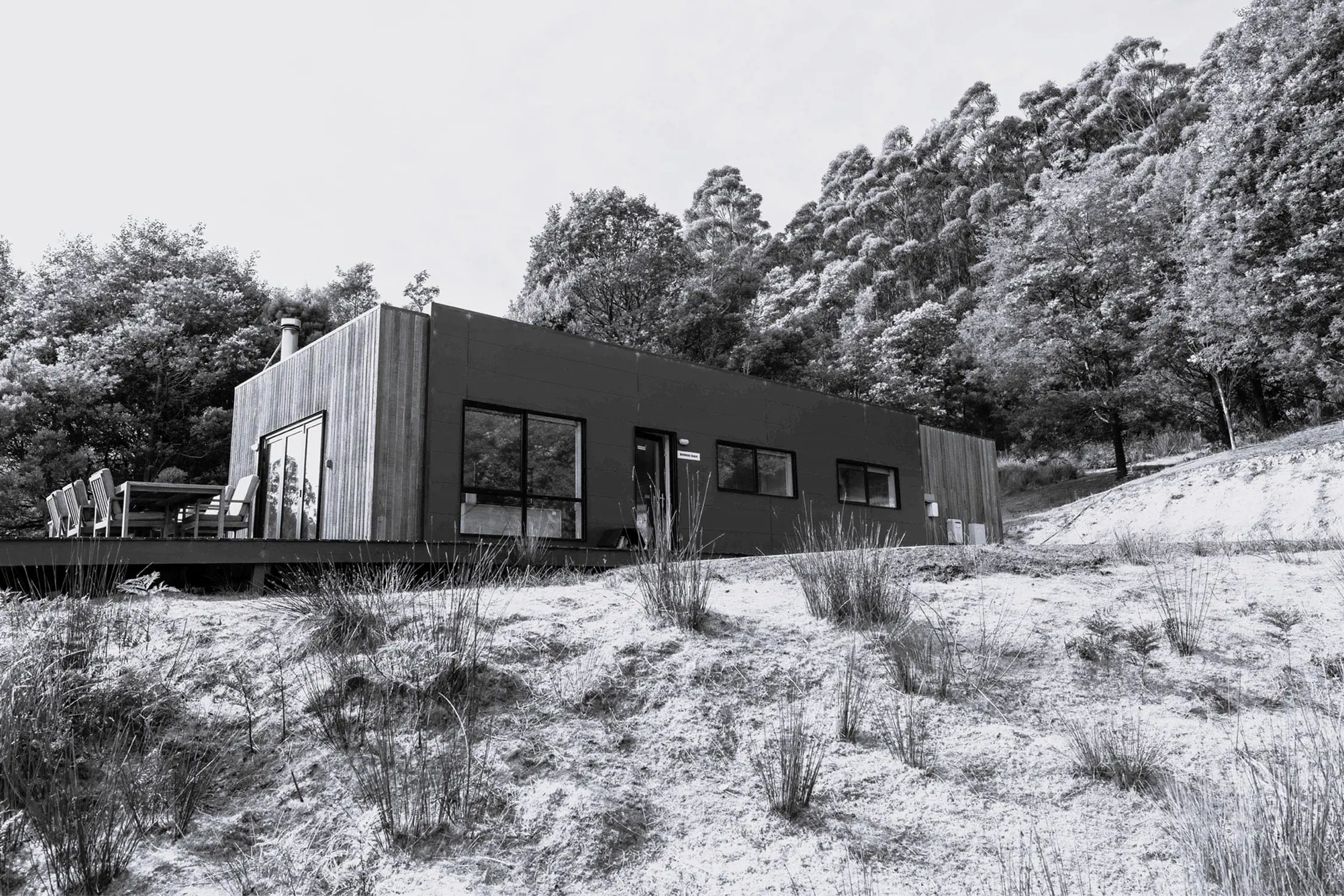 A modern rectangular cabin or house situated on a hill with surrounding trees and sparse vegetation.