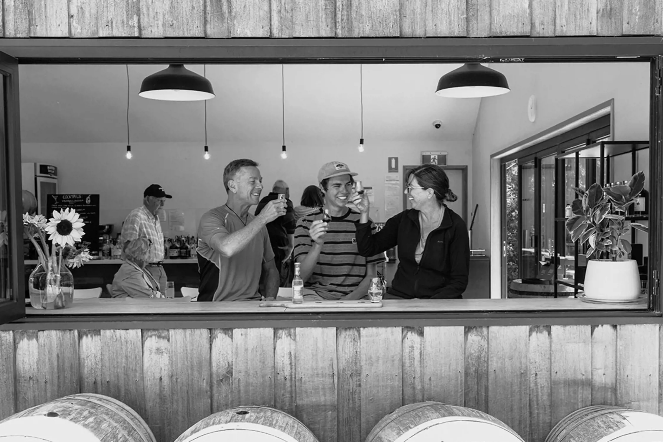 Three people sitting inside a café or bar, smiling and toasting with small glasses, visible through a wide open window.