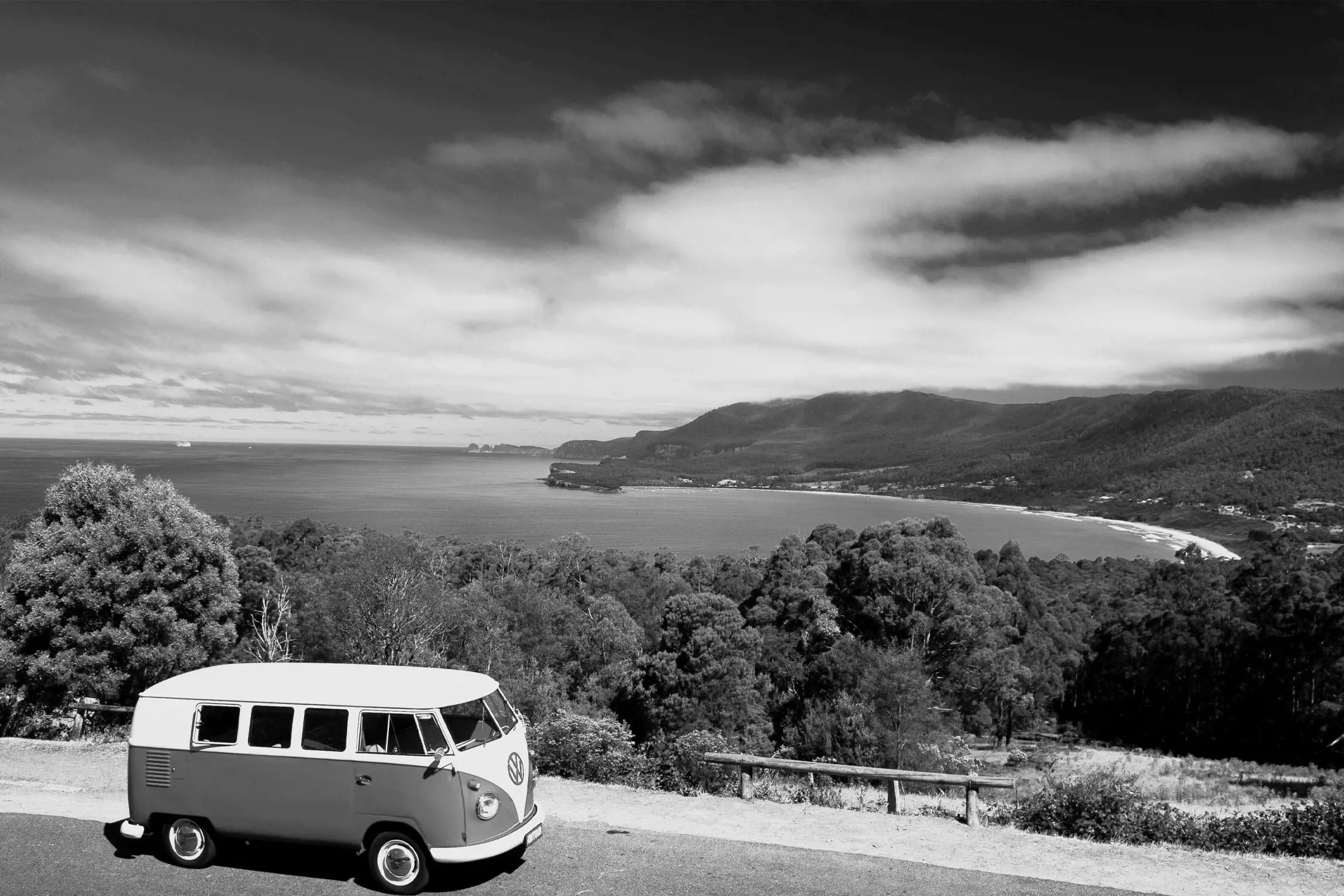 A vintage Volkswagen van parked on a roadside with a scenic coastal view showing a bay, hills, and trees in the background.