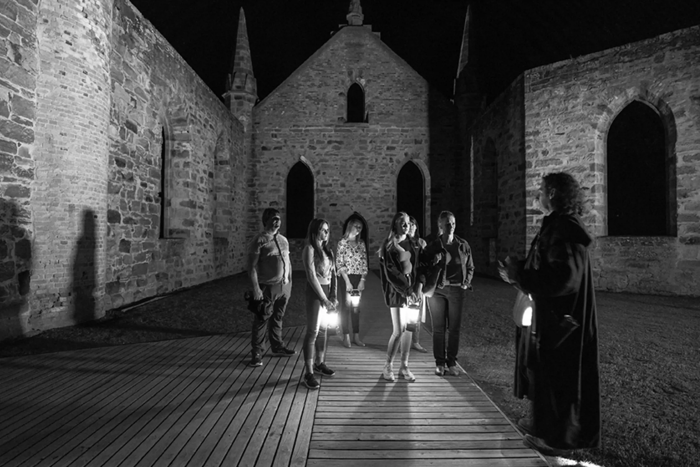 A group of people holding lanterns visiting the ruins of an old stone building at night, listening to a guide.