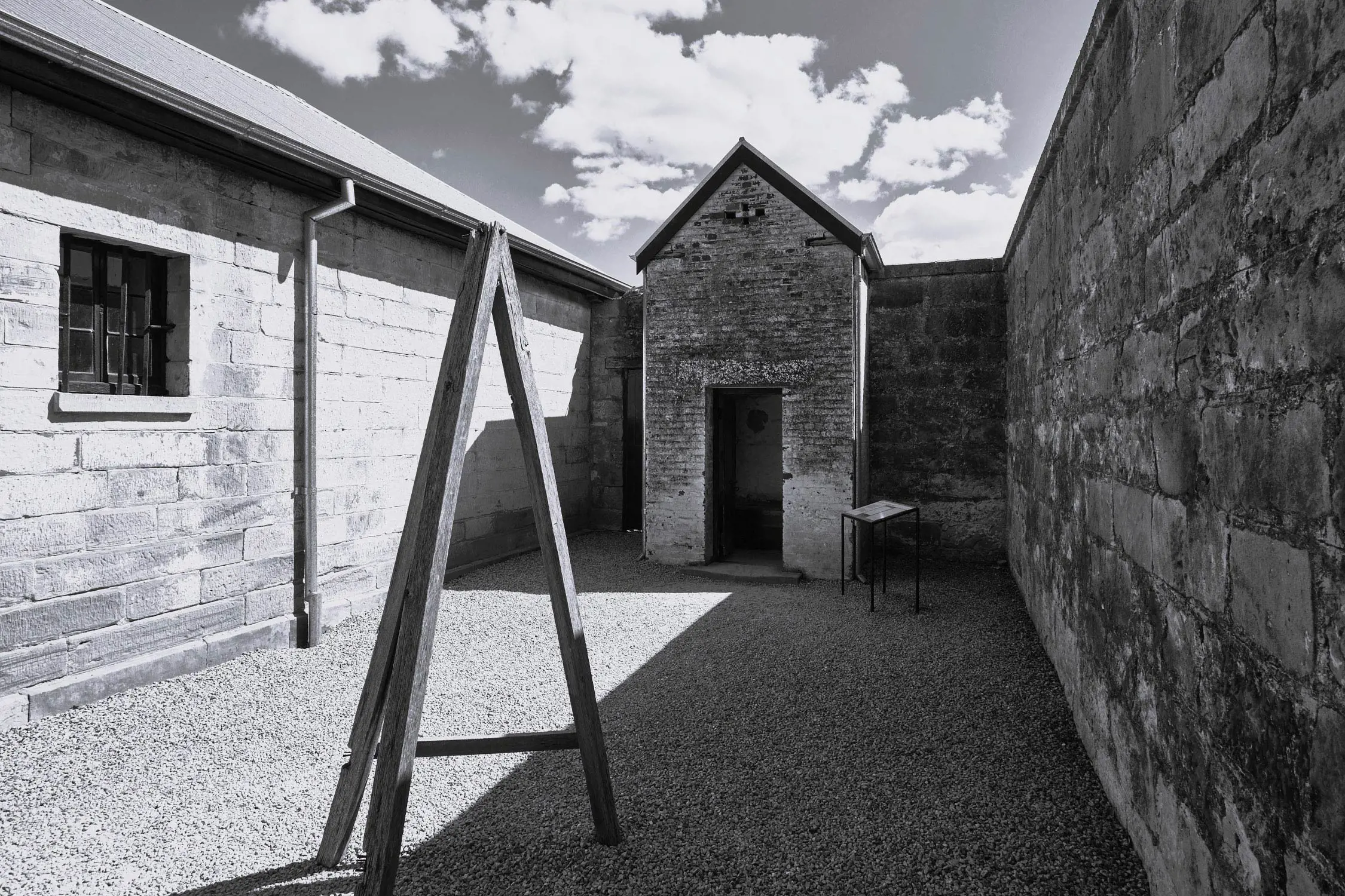 An enclosed courtyard area of a federation gaol with gravel ground and stone walls, including a small brick structure and a wooden A-frame.