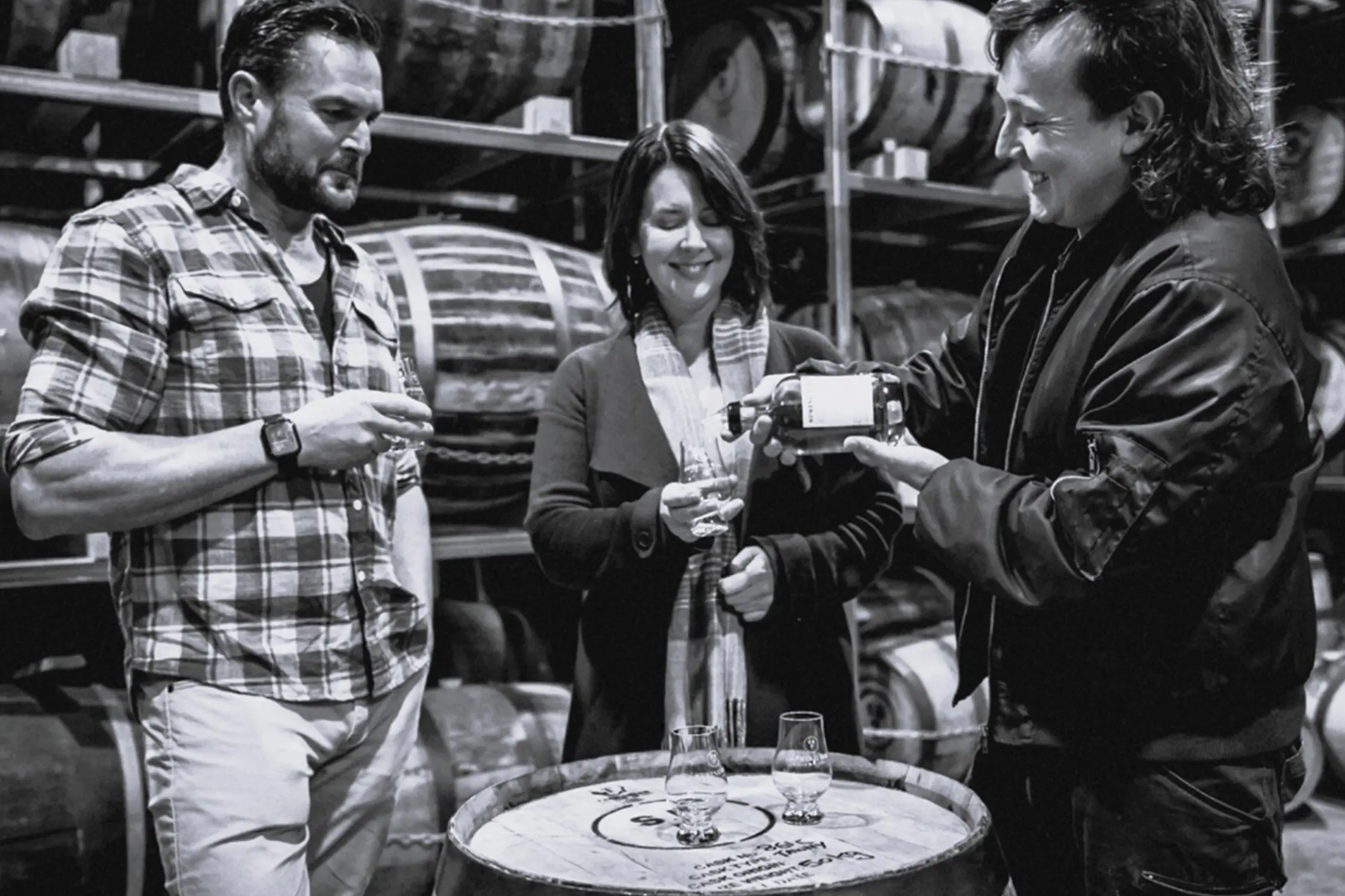 Three people in a distillery warehouse standing around a wooden barrel, one person pouring whiskey into a glass for another.