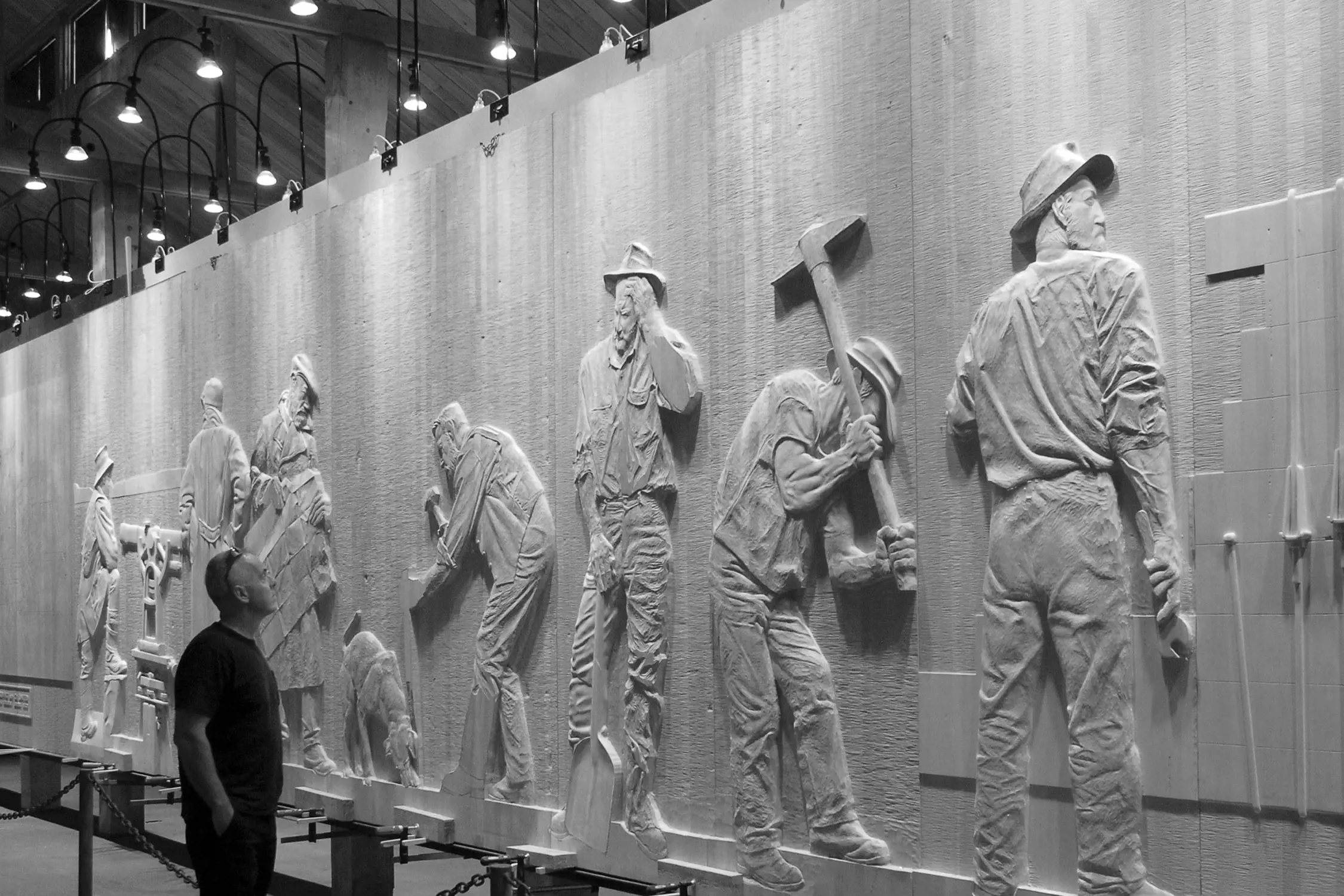 Black and white photo of a man observing a large sculptural relief mural featuring several male workers with tools. The scene is indoors with overhead lighting.