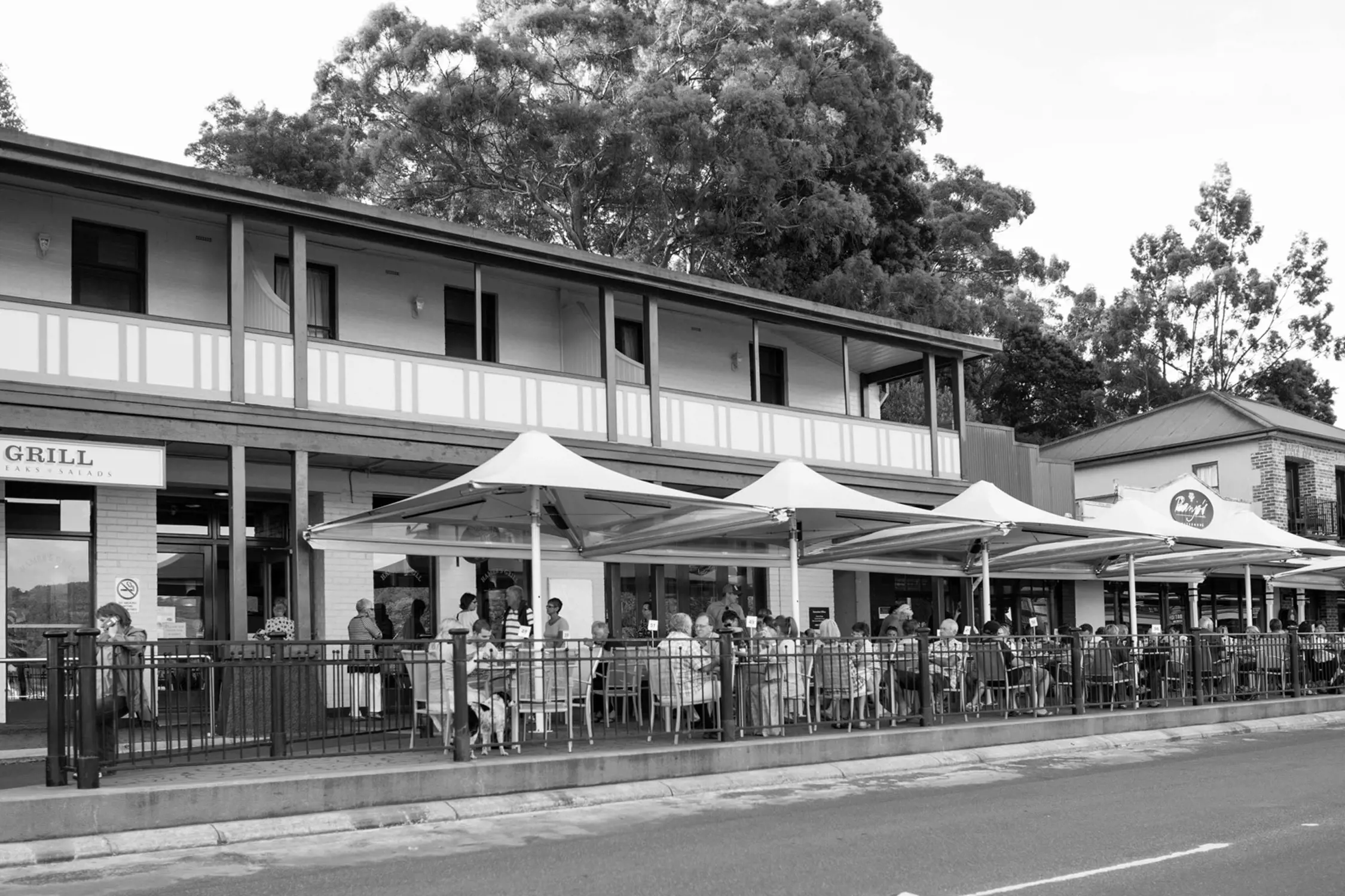 Street view of a row of buildings with outdoor seating at cafés and restaurants, many people dining or socializing under umbrellas, a tree-lined street in the foreground.