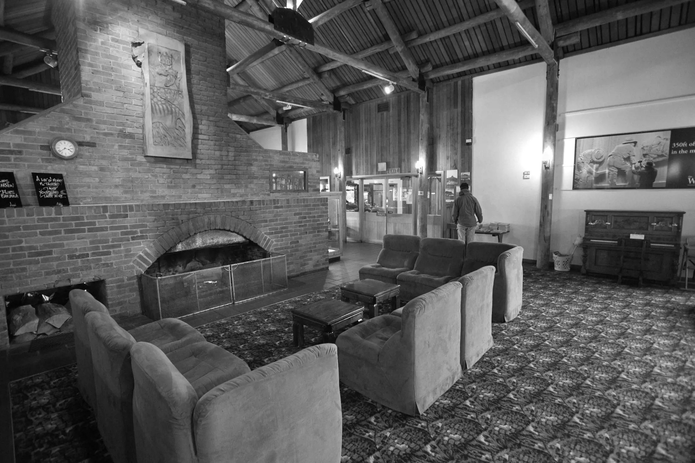 Interior of a rustic lodge or cabin with a large brick fireplace, several cushioned chairs arranged around small tables, a patterned carpet, wooden beams on the ceiling, and a person walking near the entrance doors.