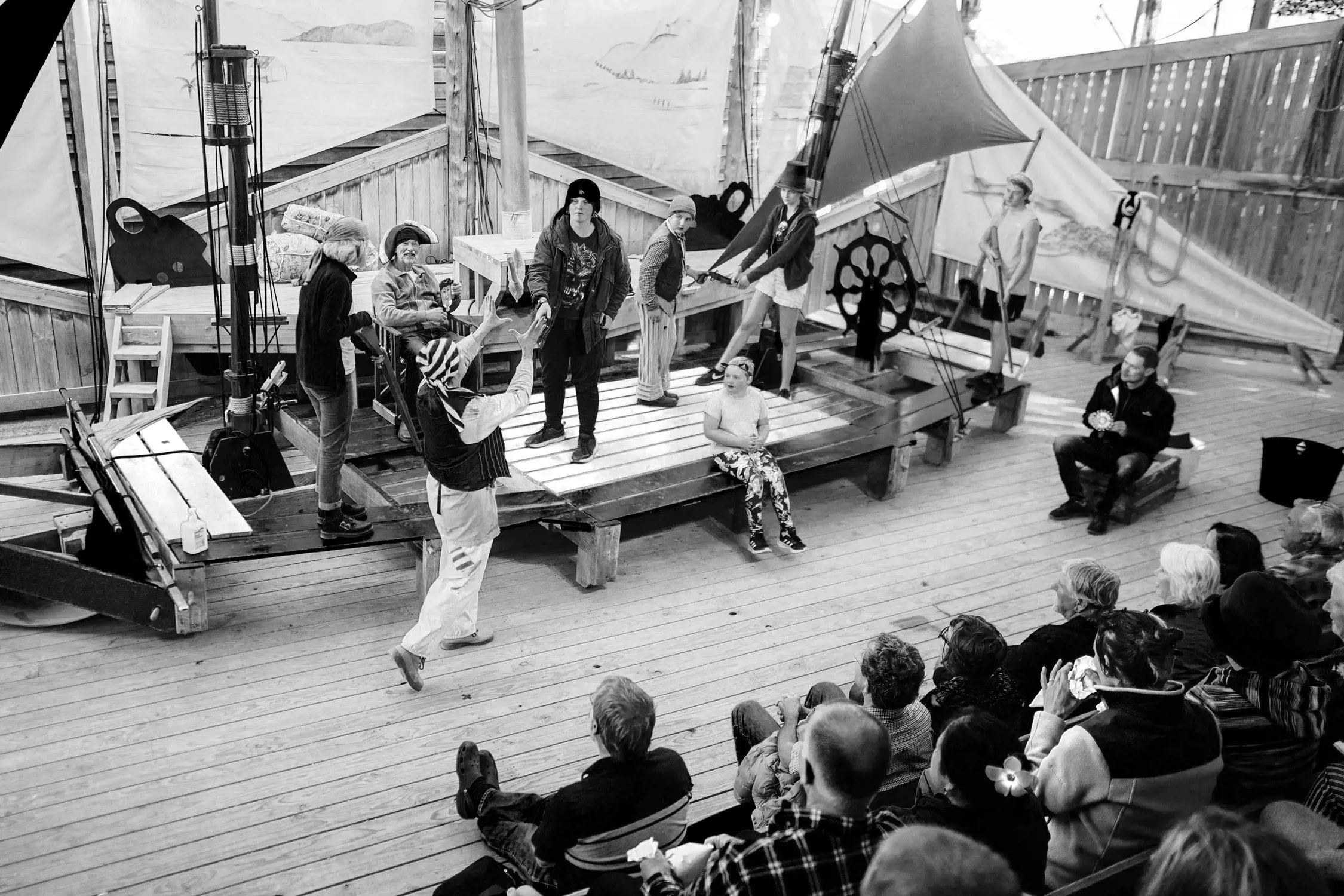 Black and white scene of a stage play or reenactment with actors in costumes performing on a wooden deck resembling a ship, an audience seated in front watching attentively.