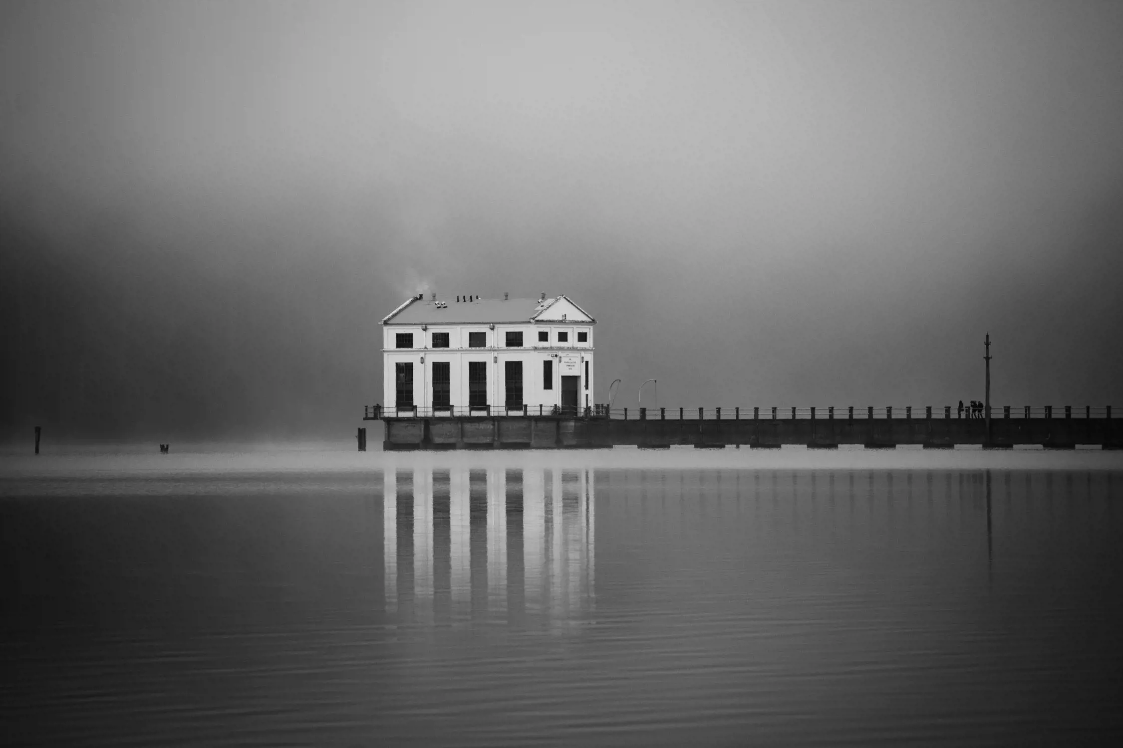 A misty dawn or dusk view of a white building on a pier extending into calm water with reflections.