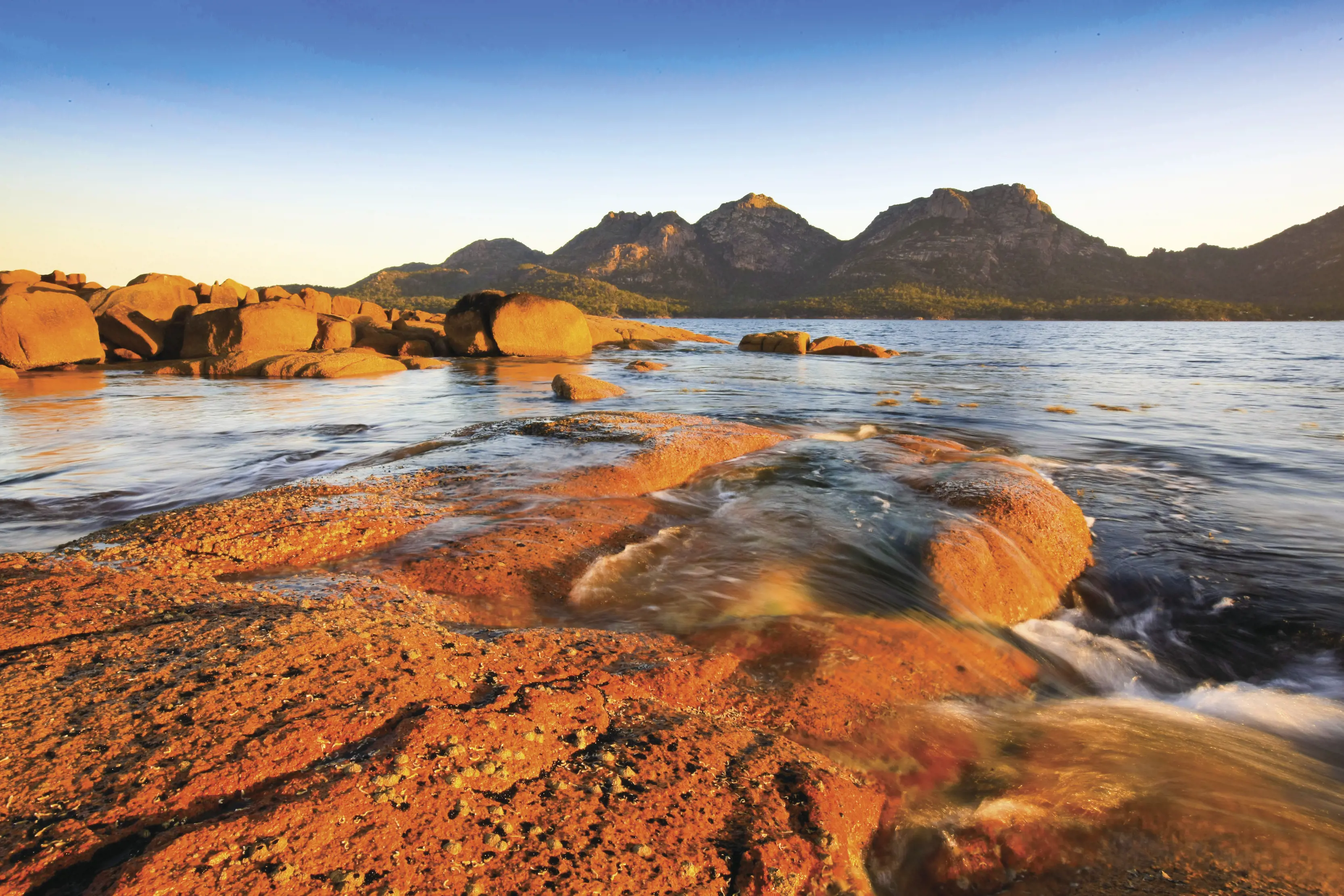View of the The Hazards in Coles Bay. The rocks shine red as the sun sets over the blue lake.