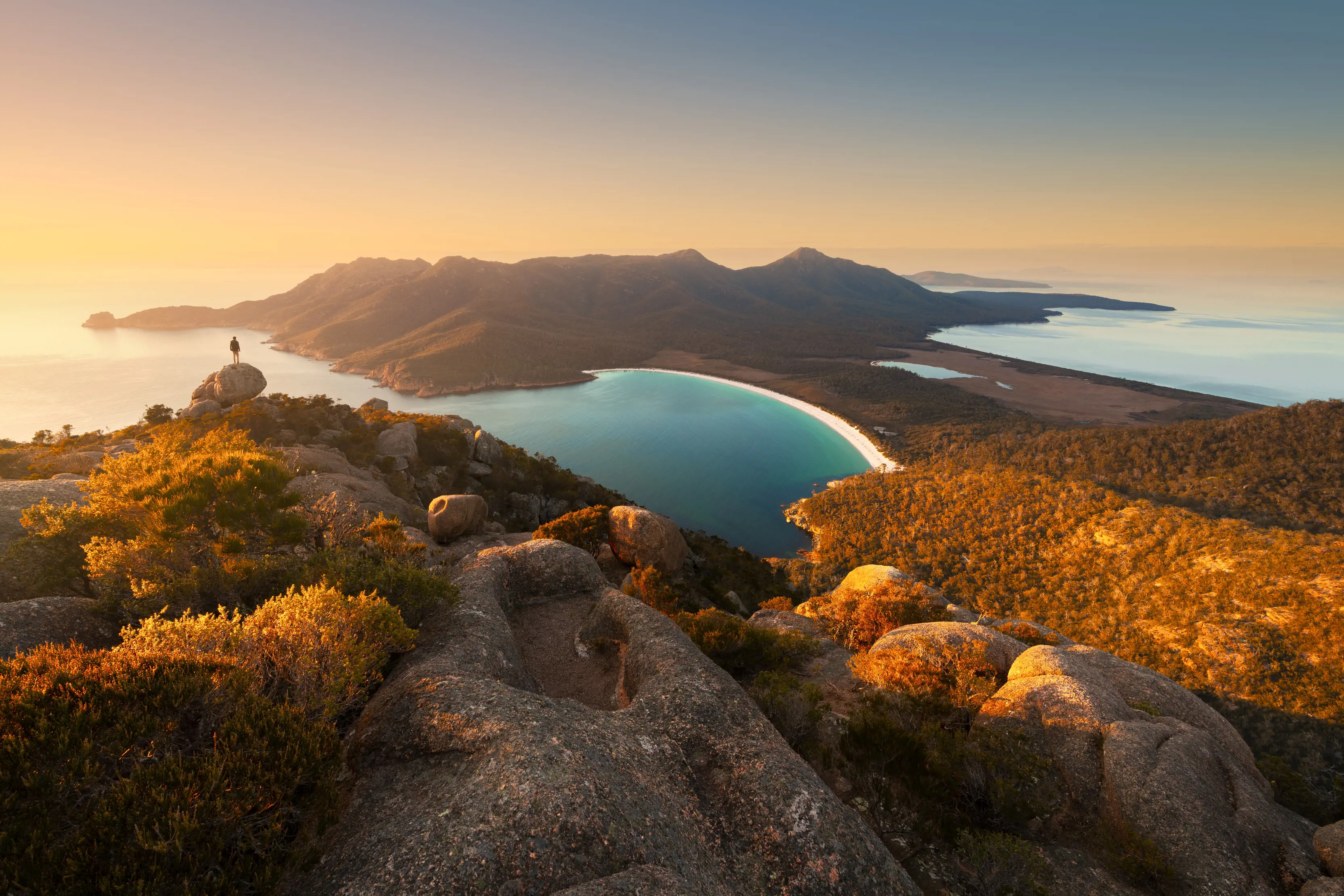 Aerial shot of Wineglass Bay, Freycinet National Park. A man standing on the cliff, looking out onto the water.