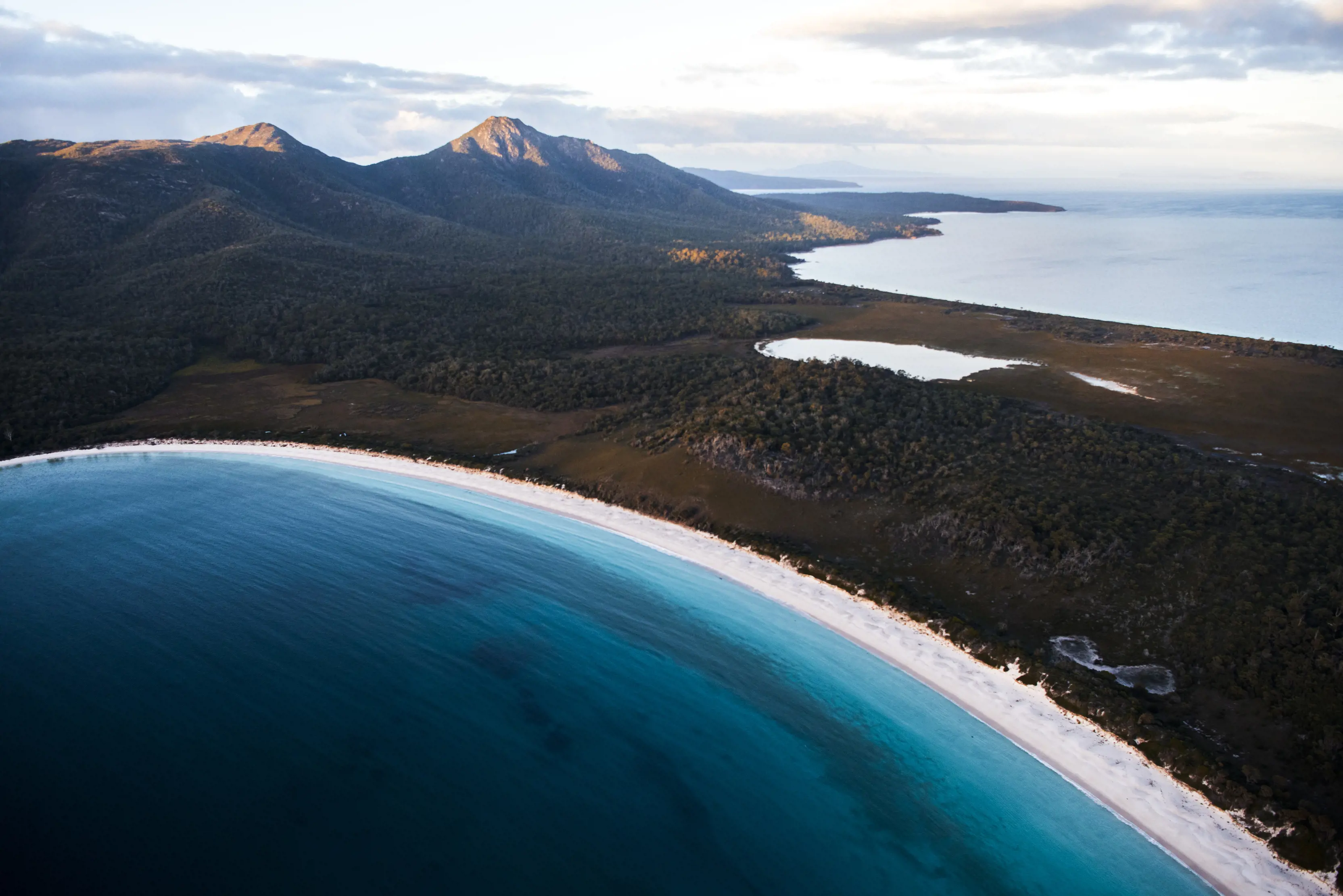 Aerial view of Wineglass Bay beach. Dark blue waters. Mountains in the background and another lake.
