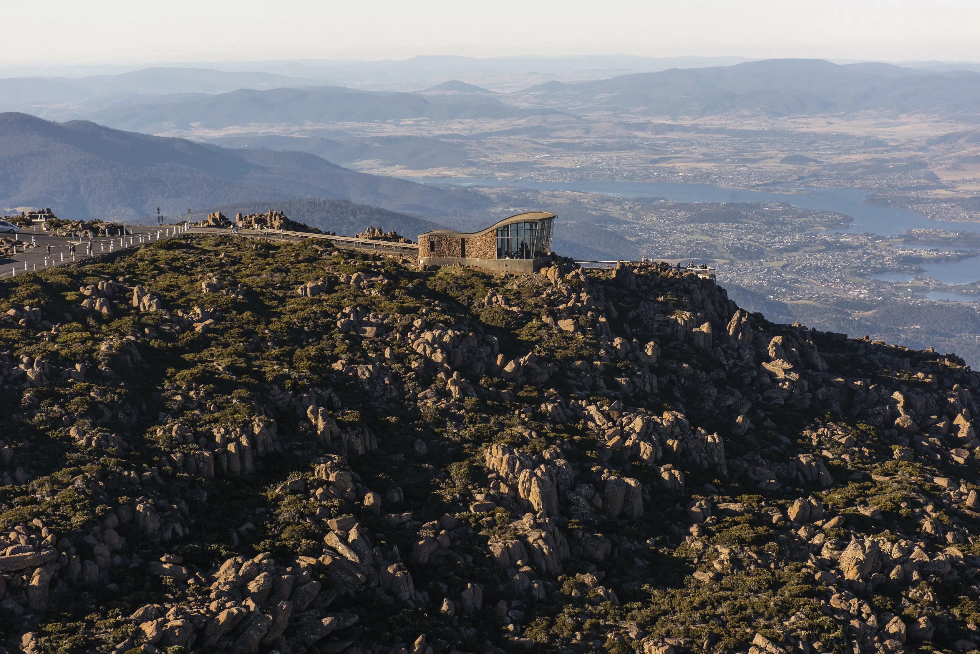 Incredible aerial view of Kunanyi / Mt Wellington with a lookout building at the top. Sun hits the rocks for a dramatic and vibrant scene.