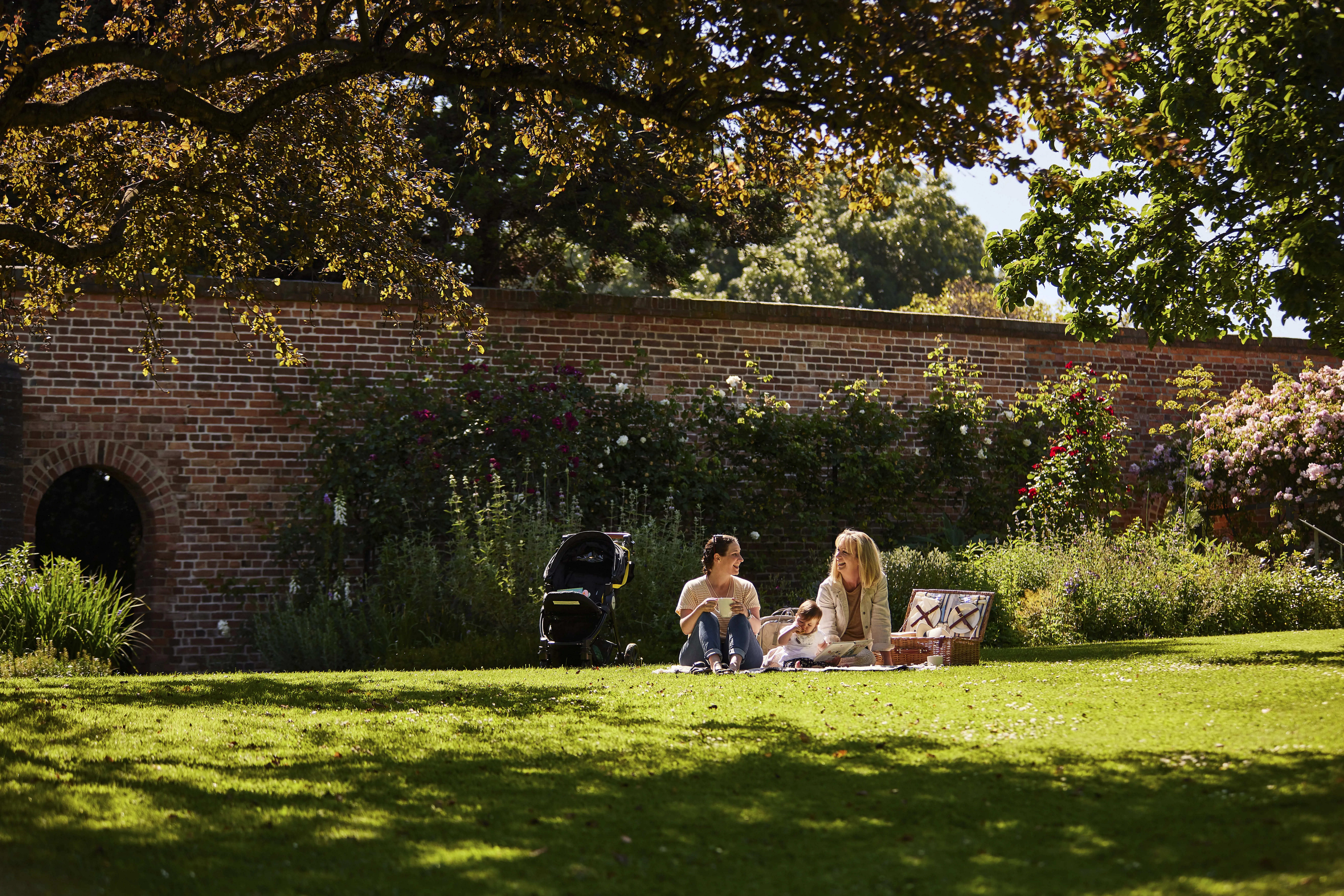 A family picnics in the sun on the grass in front of a long, tall red brick wall.