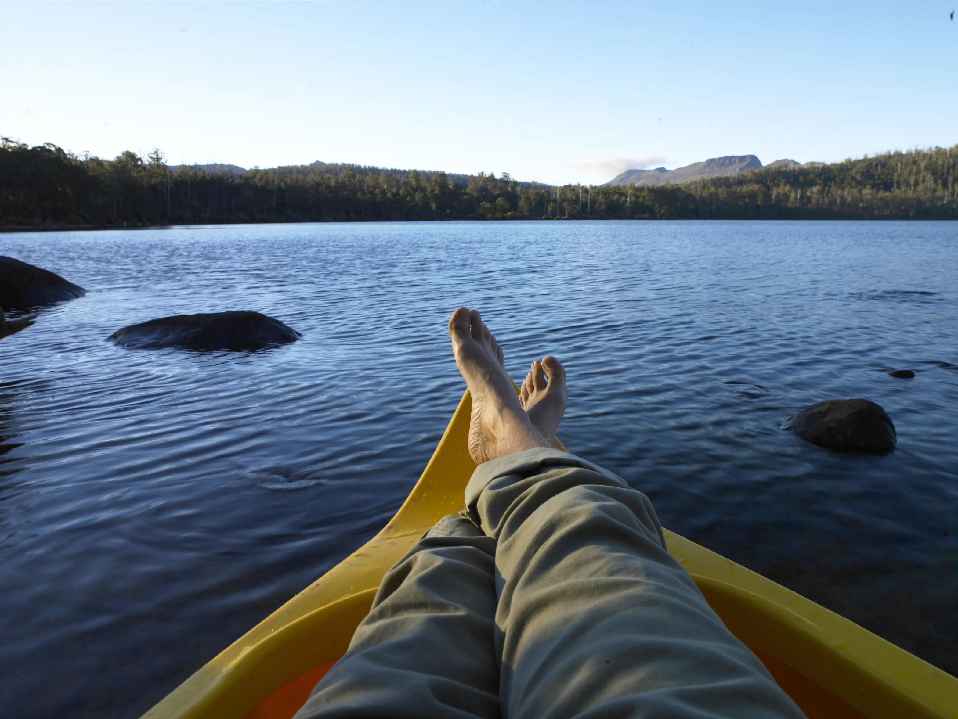 Taken from the view of the traveller, a man relaxes crossing his legs on a yellow kayak on Lake St Clair.