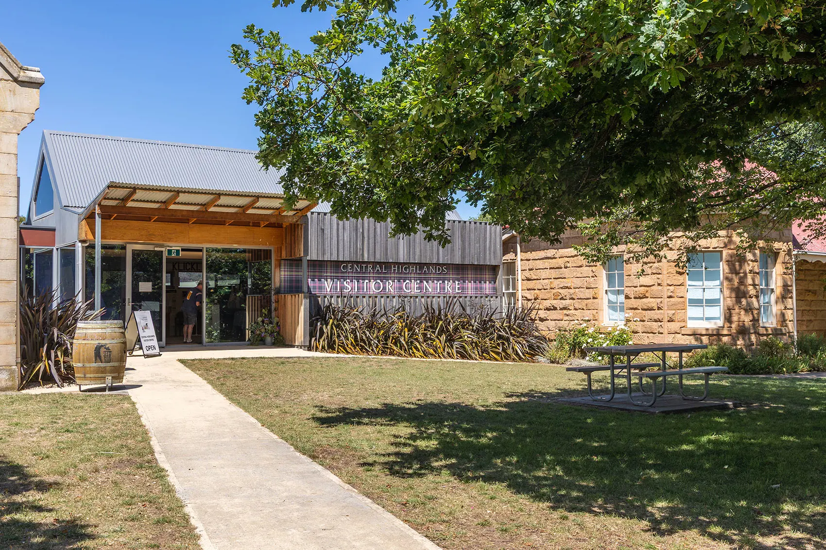 A pathway across lawn leads to a mix of old and new buildings clustered together. The sign, on a tartan backing, reads "Central Highlands Visitor Centre".