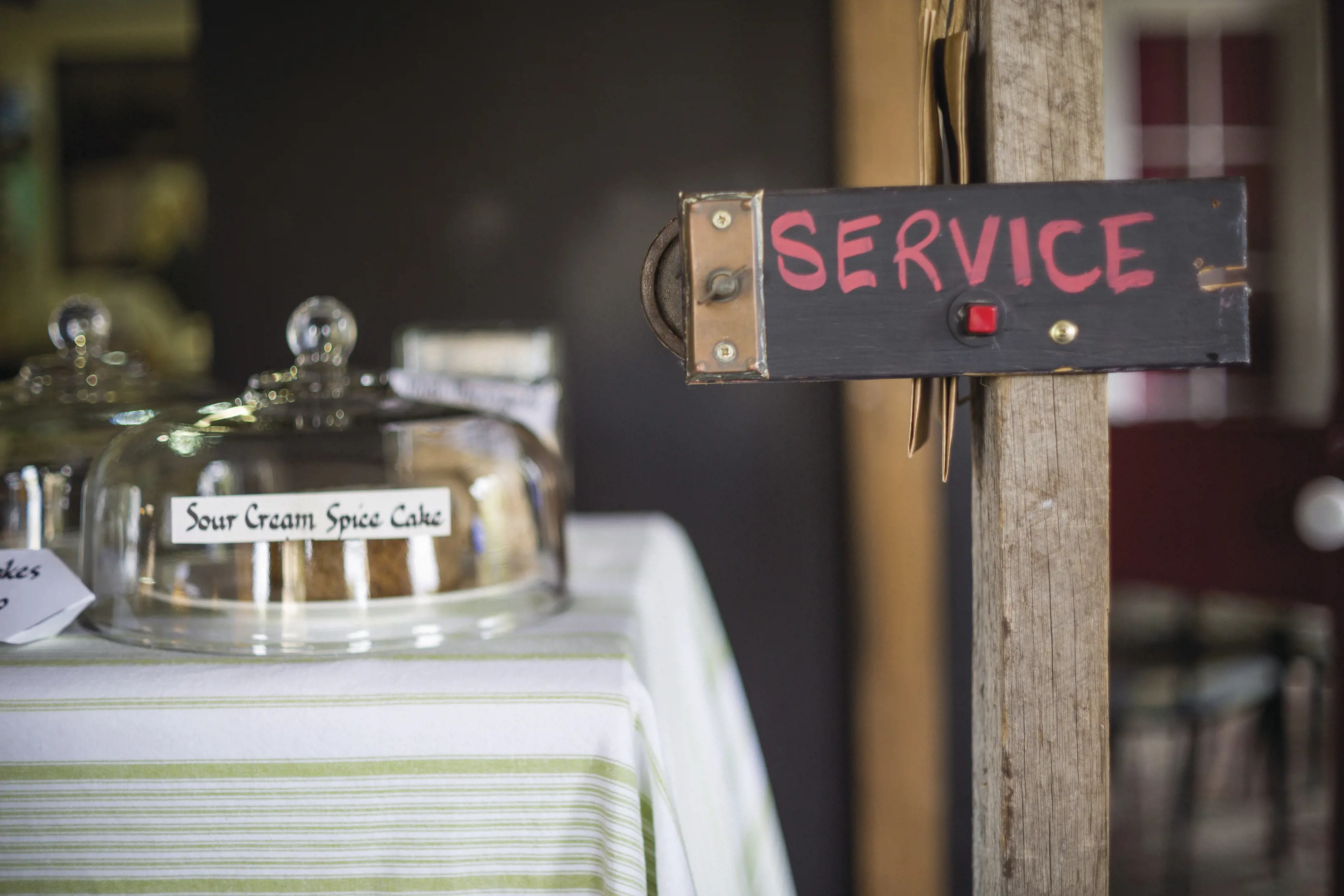 Image of Sour Cream Spice Cakes for sale under a cake cover at Jackson's Emporium, there is a service chalk sign.