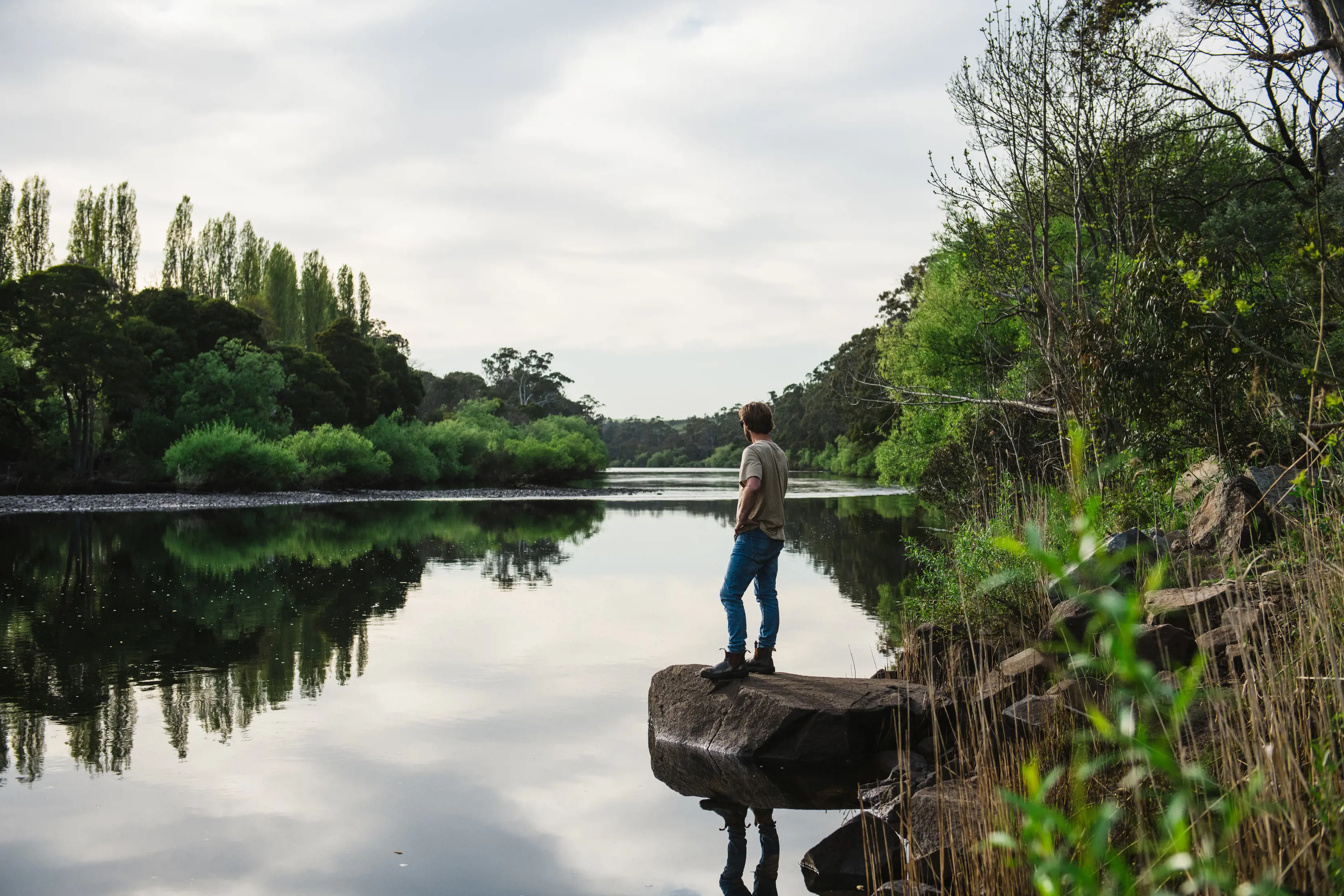 A man stands on a rock jutting out into a river. The water's surface is still and reflects the bushy banks around it.