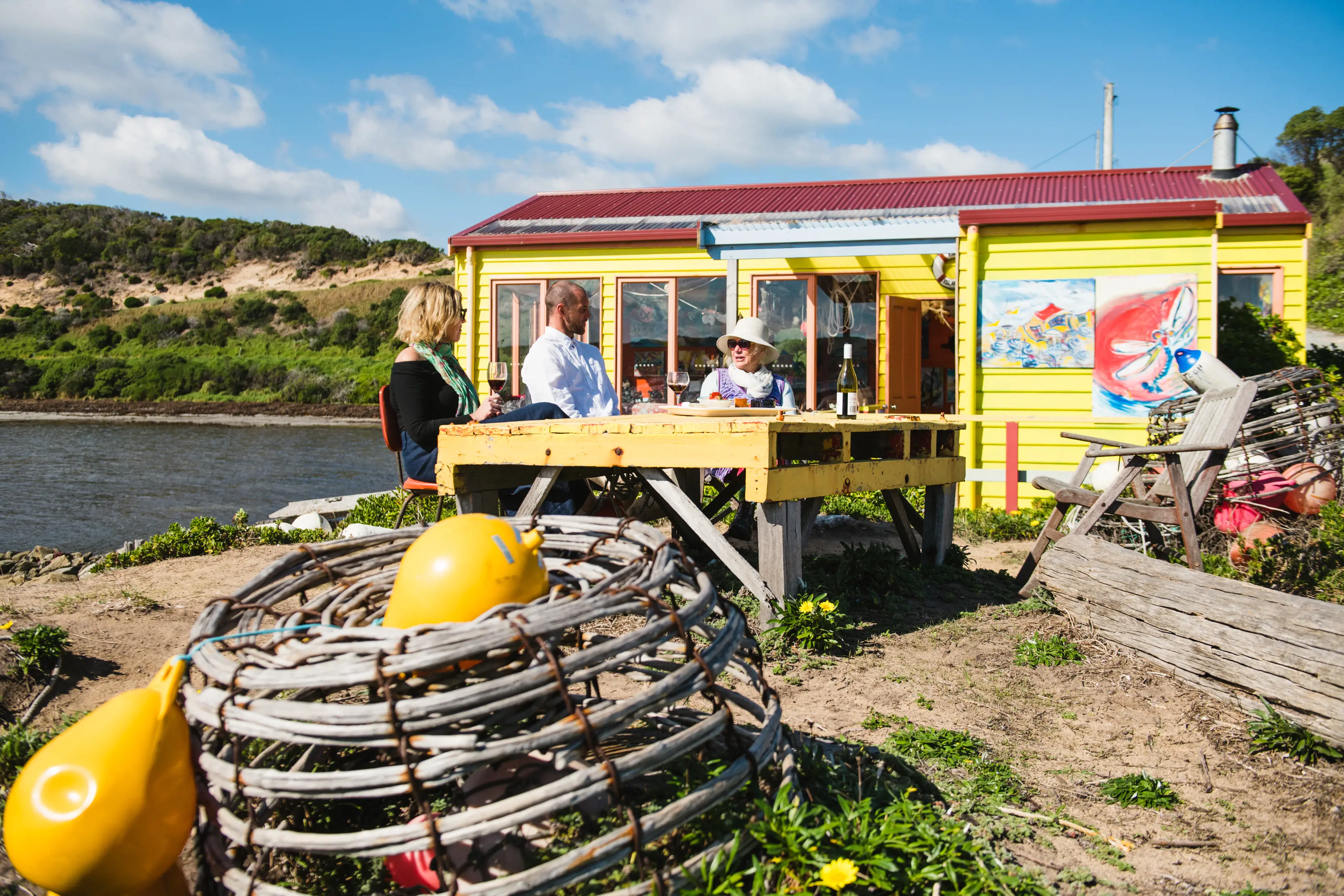 A group of three people sit outside The Boathouse - The Restaurant With No Food, drinking wine, by the water.