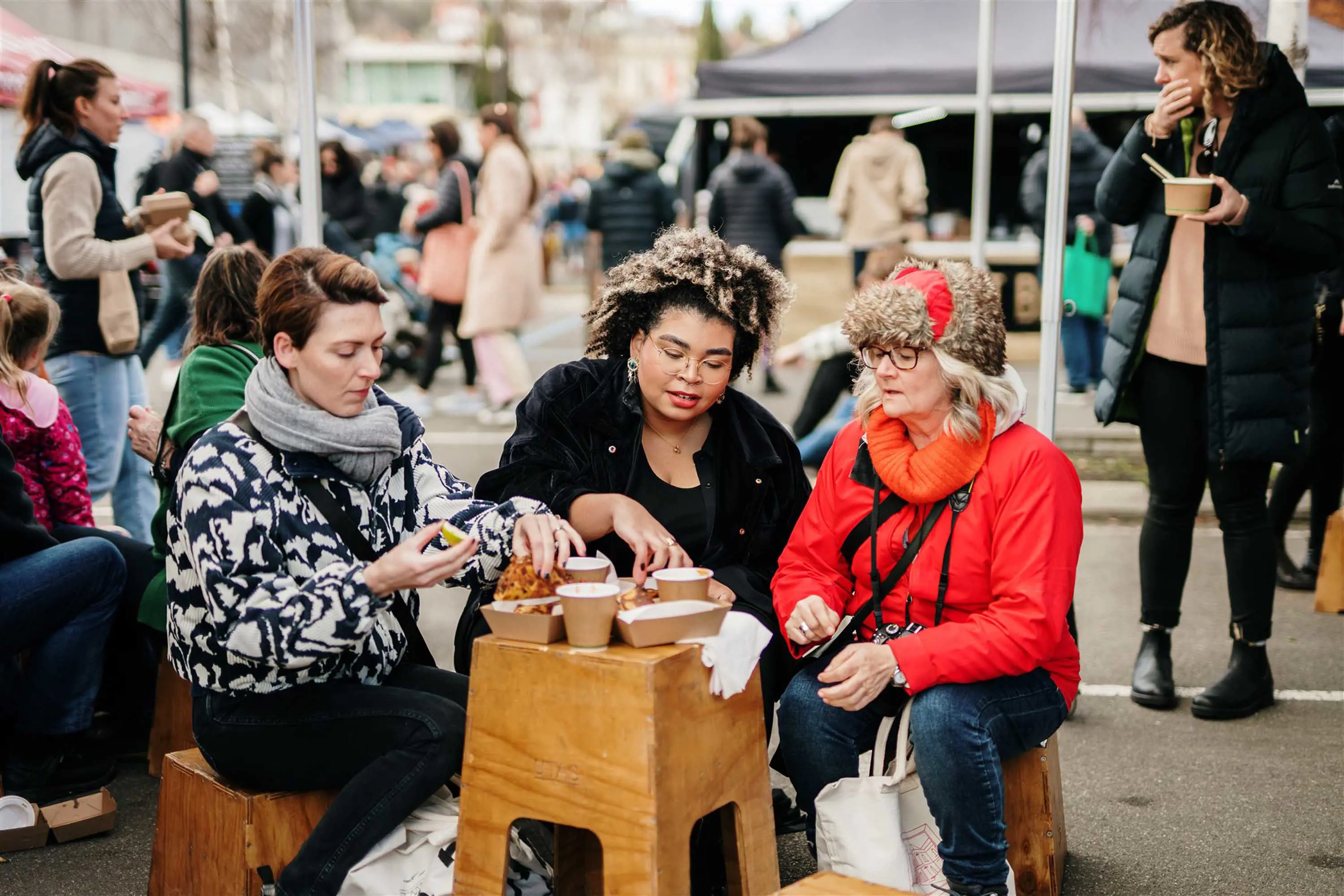 A group sit around a makeshift table, drinking coffee and eating food from market stalls.