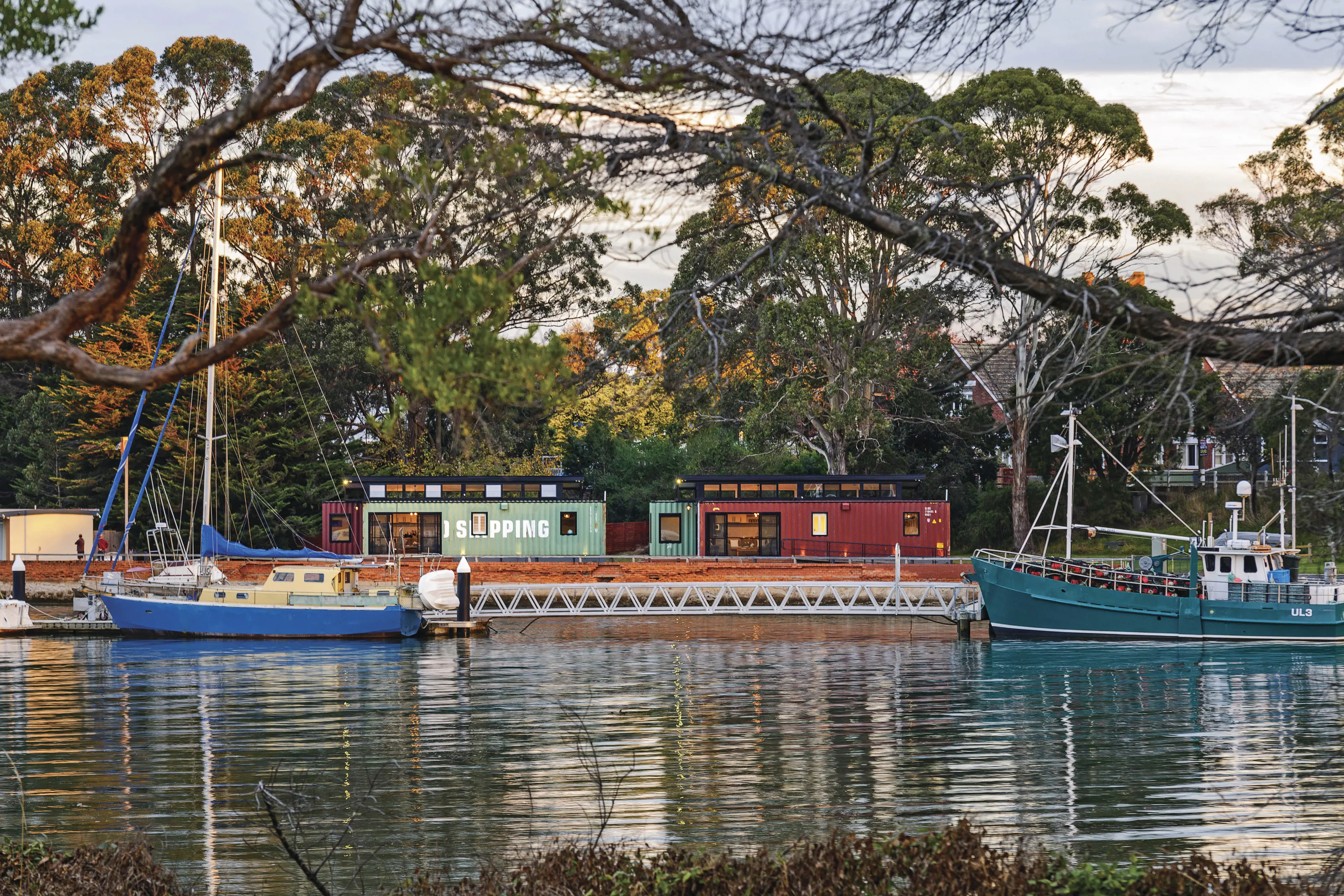 Colourful small boats are lined up moored on a river where two shipping container buildings sit on the bank.
