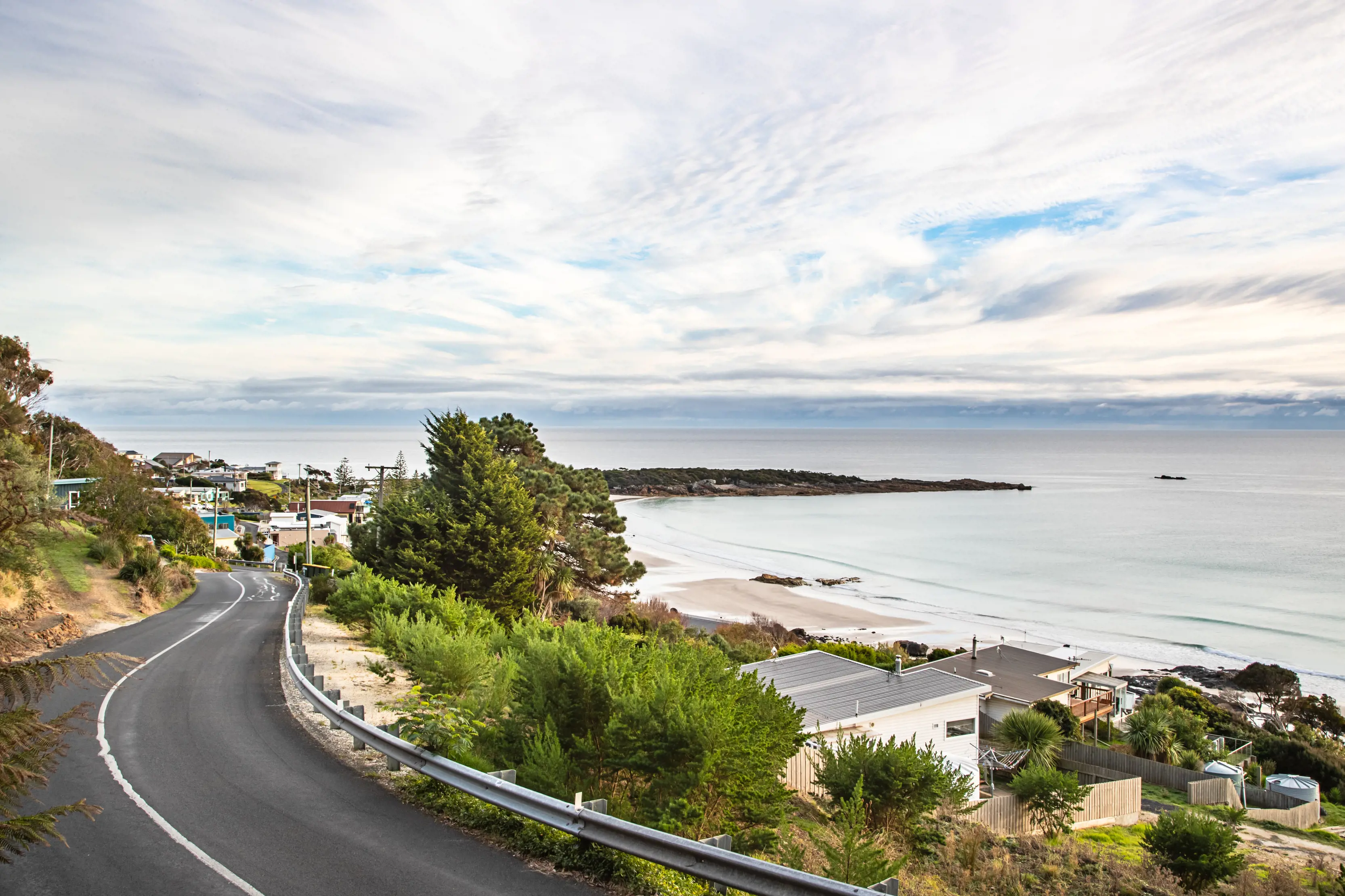 Image of a windy road into Boat Harbour on the left side of the image. Houses fill the right of the image that lead to the shoreline and ocean.