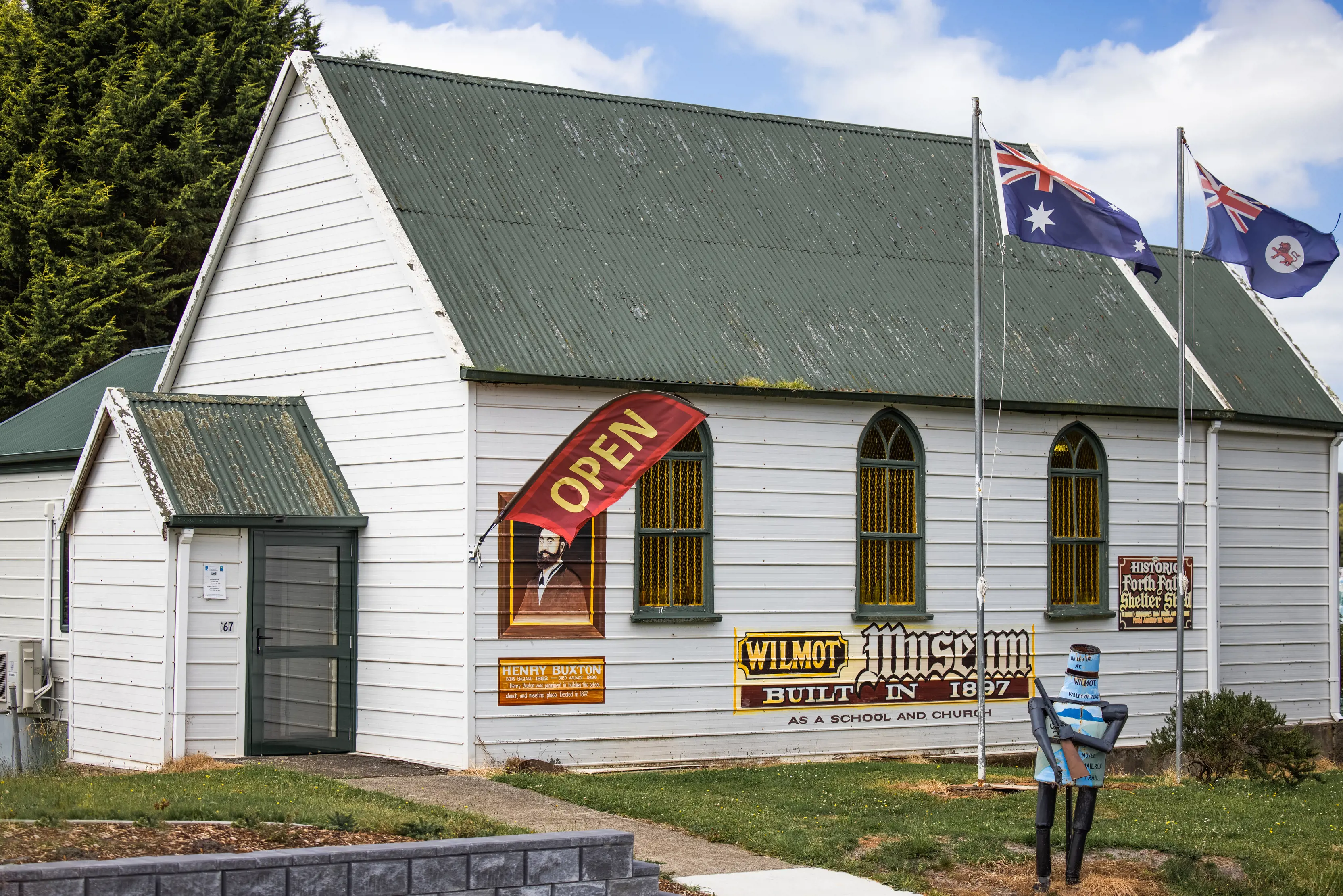 Image of the outside of the Wilmot Museum. A white building with a khaki green roof. A flag sits out the front that reads "Open".