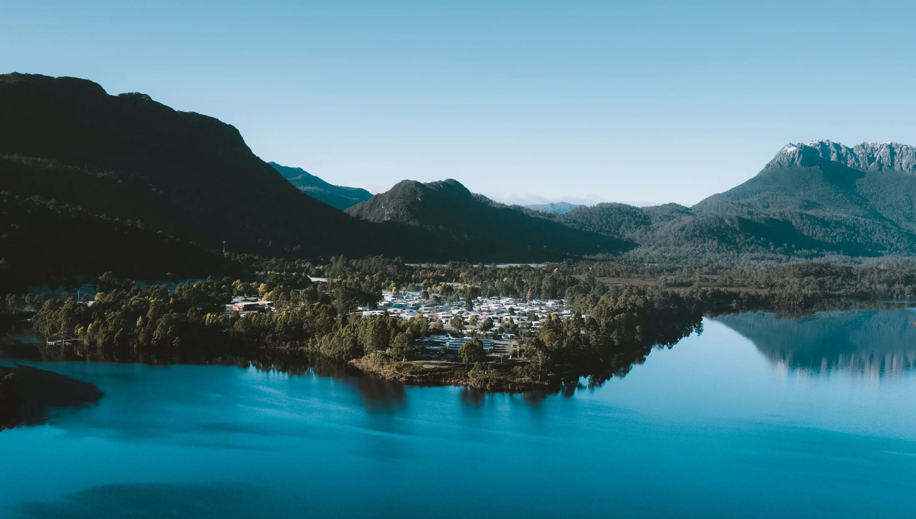 An aerial image of a small rural town, nestled amongst trees on the edge of a lake. Mountains surround the town in the background, rising sharply with snow on their peaks.