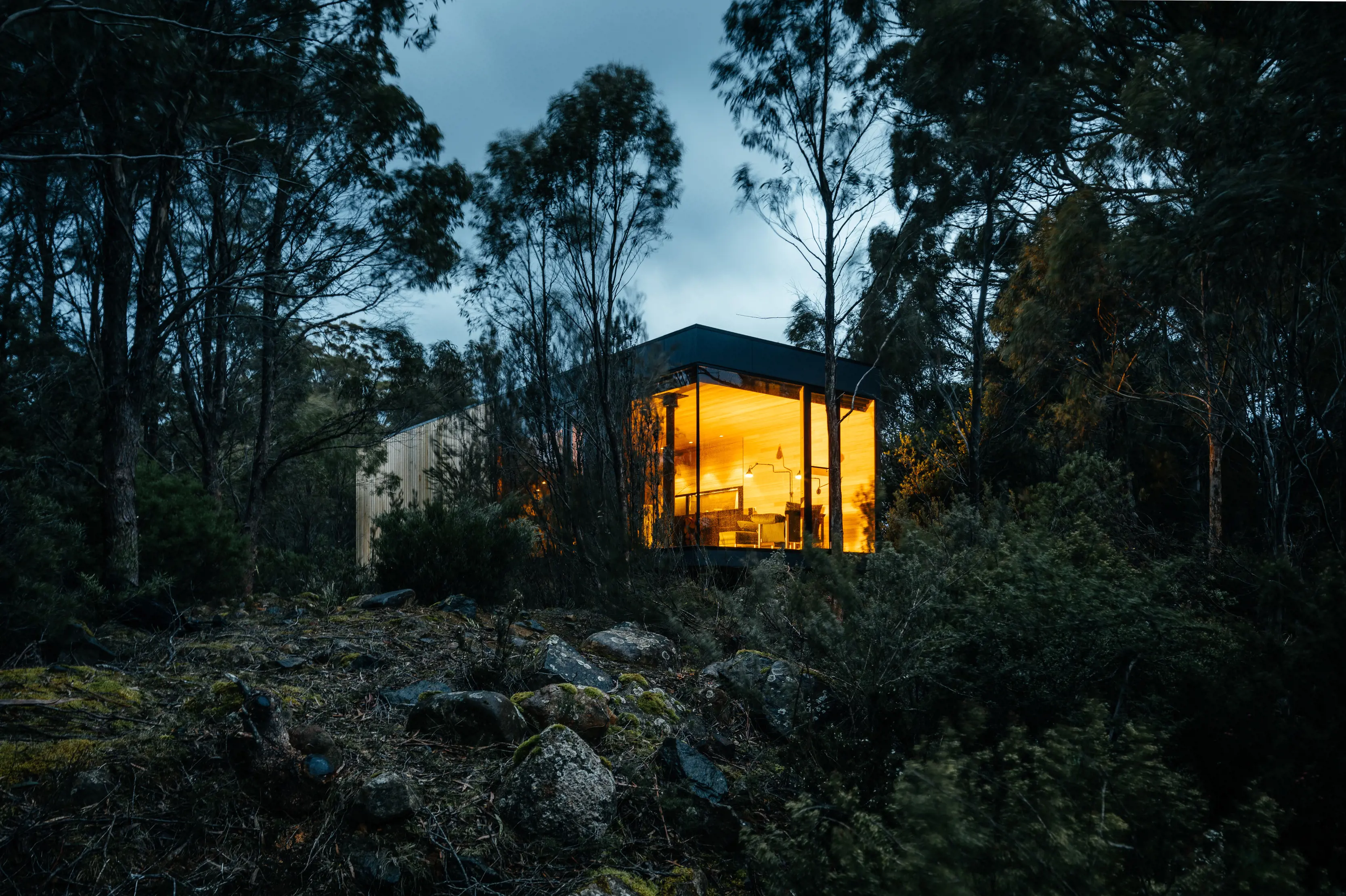 Nightfall shot of The Retreat in Pumphouse Point. Surrounded by tall trees and the view of the cabin's glowing light shining through the darkness.