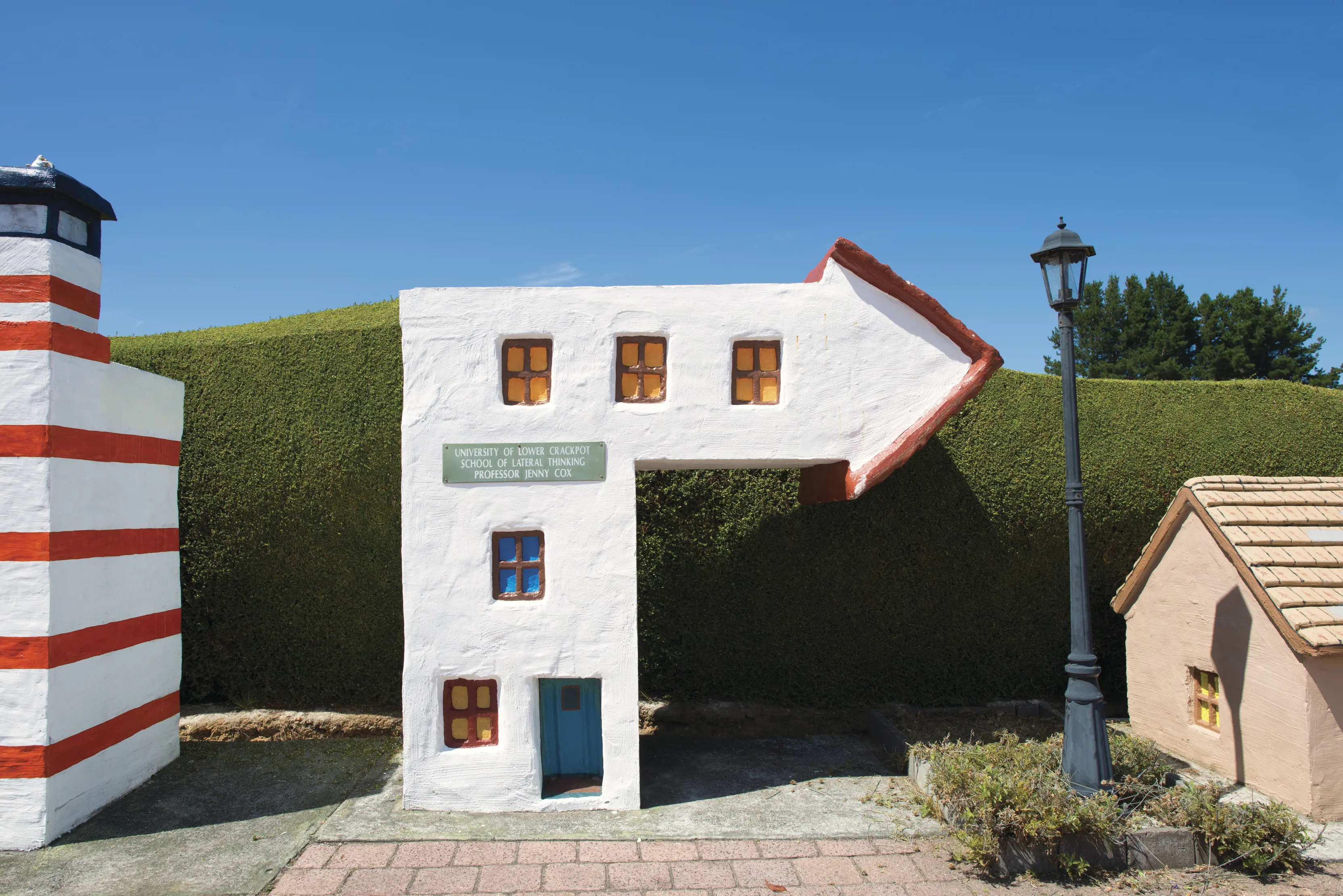 A small sculptural house, shaped like an arrow pointing right. A large hedge frames the background.