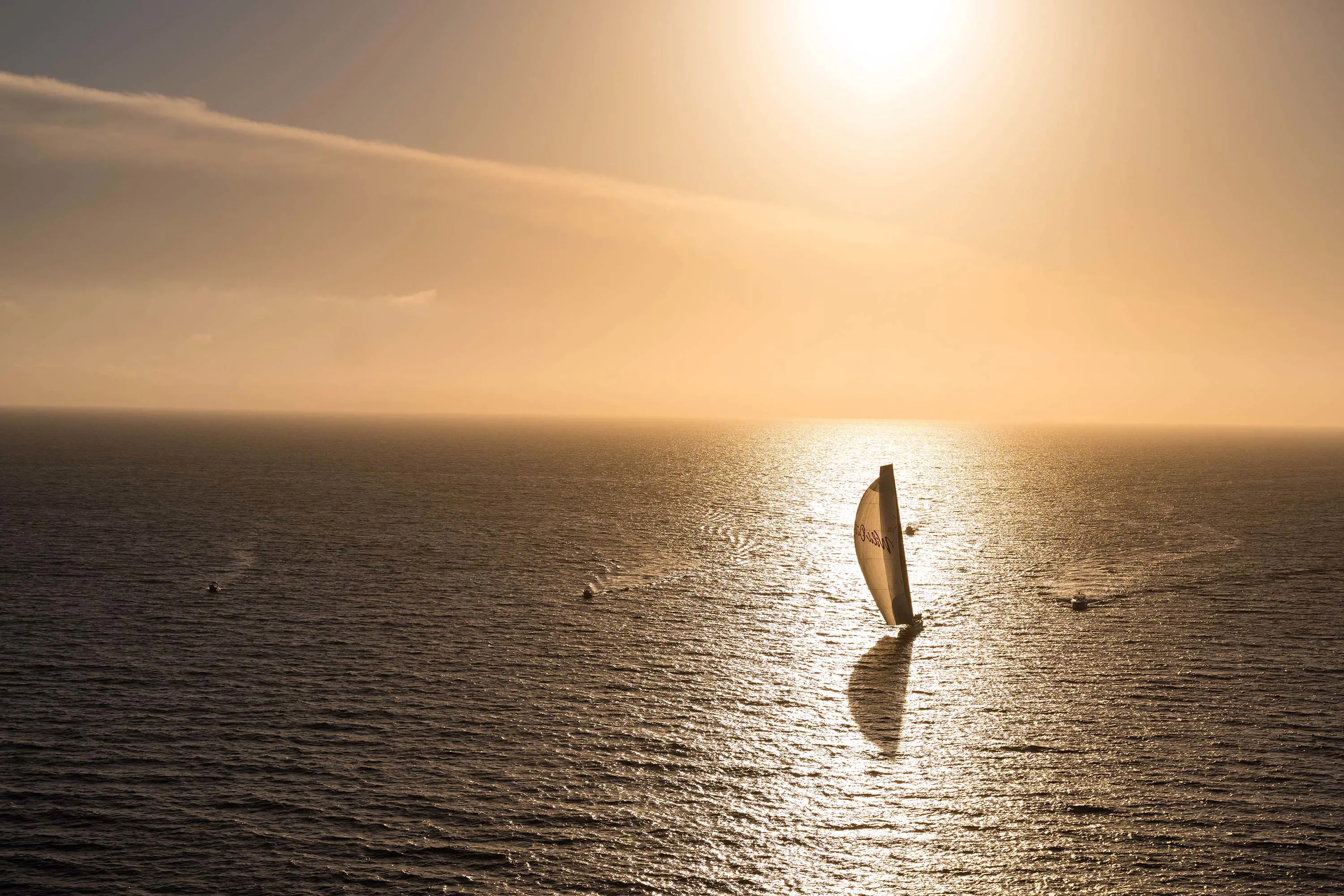 A lone sailing yacht on the ocean, with three little support boats nearby. The yacht is framed by the sun's reflection on the water.