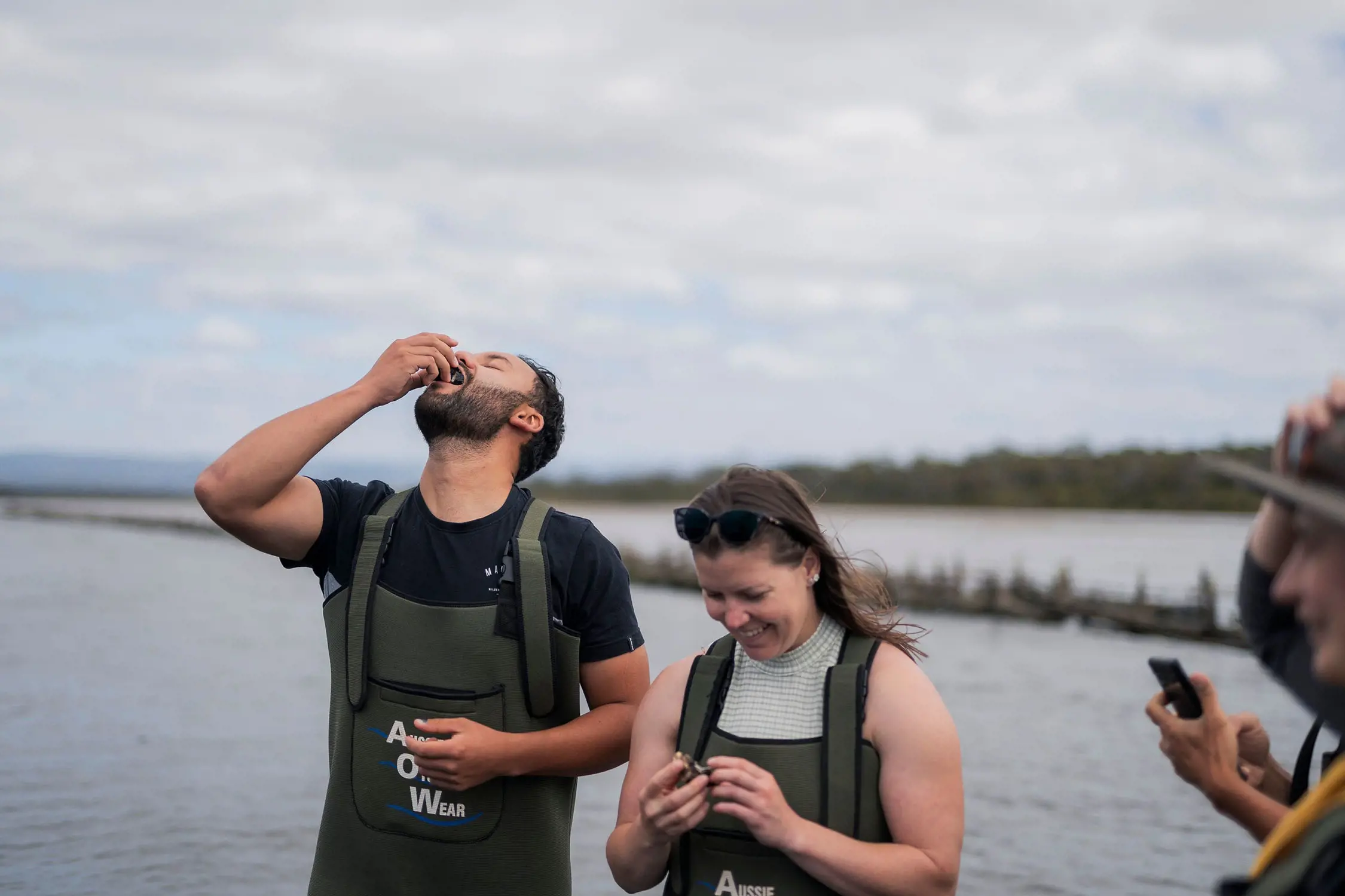 Two people wearing wading overalls are slurping oysters straight from their shells. In the background are the still waters of an ocean's inlet.