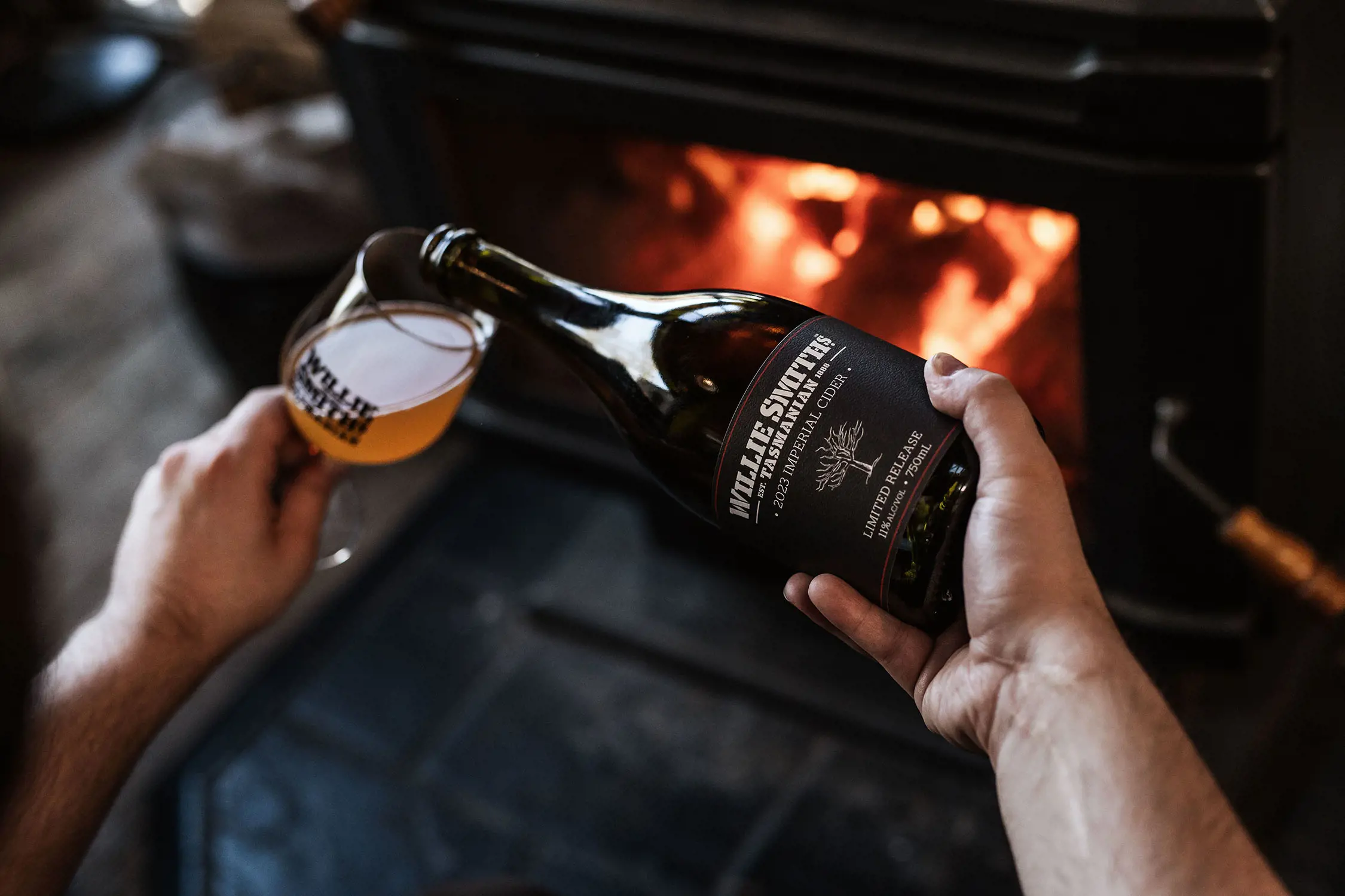 In front of a oaring fireplace, a person pours cider from a bottle into a glass. Both are labelled 'Willie Smith's'.