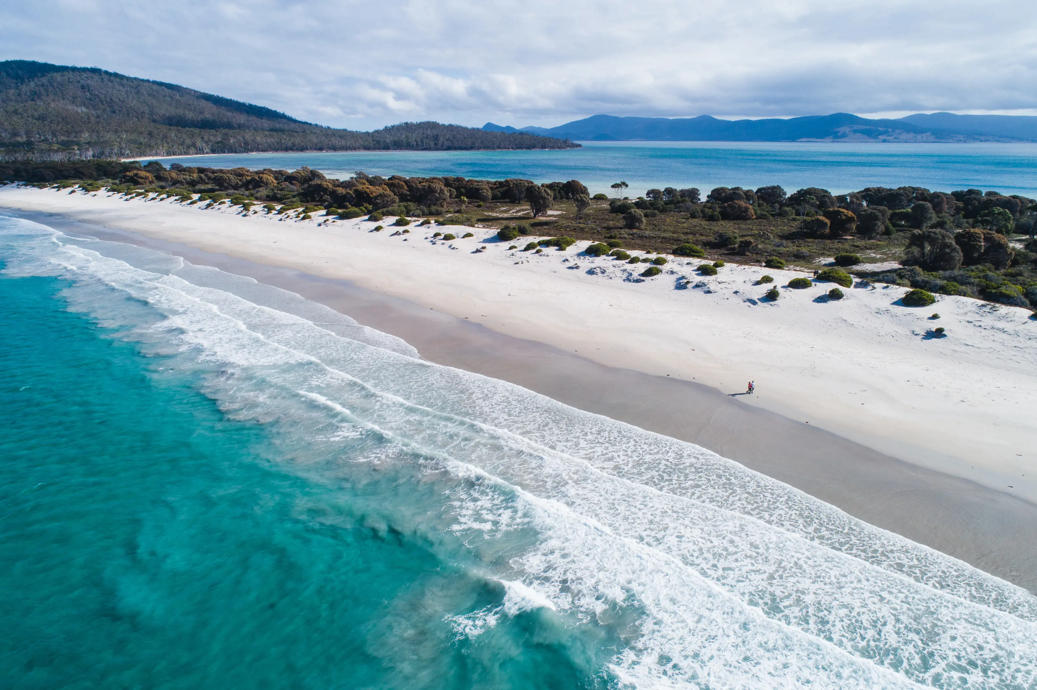 An aerial view of a picturesque beach, with bright teal blue water, waves rolling in and white sand.