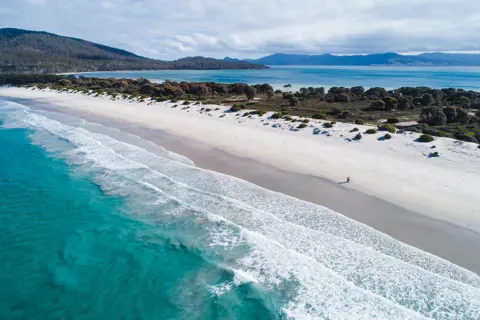 An aerial view of a picturesque beach, with bright teal blue water, waves rolling in and white sand.