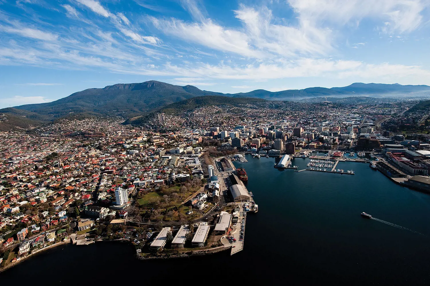 An aerial view of Hobart's waterfront and wharf, city and suburbs beyond. The skyline is dotted with mountain ranges.