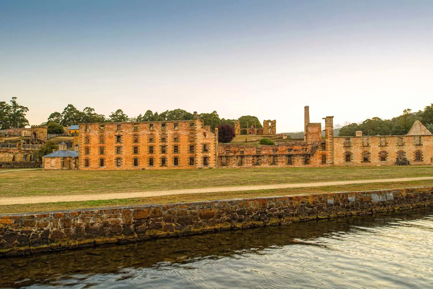 A row of heritage brick built, partly ruined buildings alongside neat lawn and a canal.