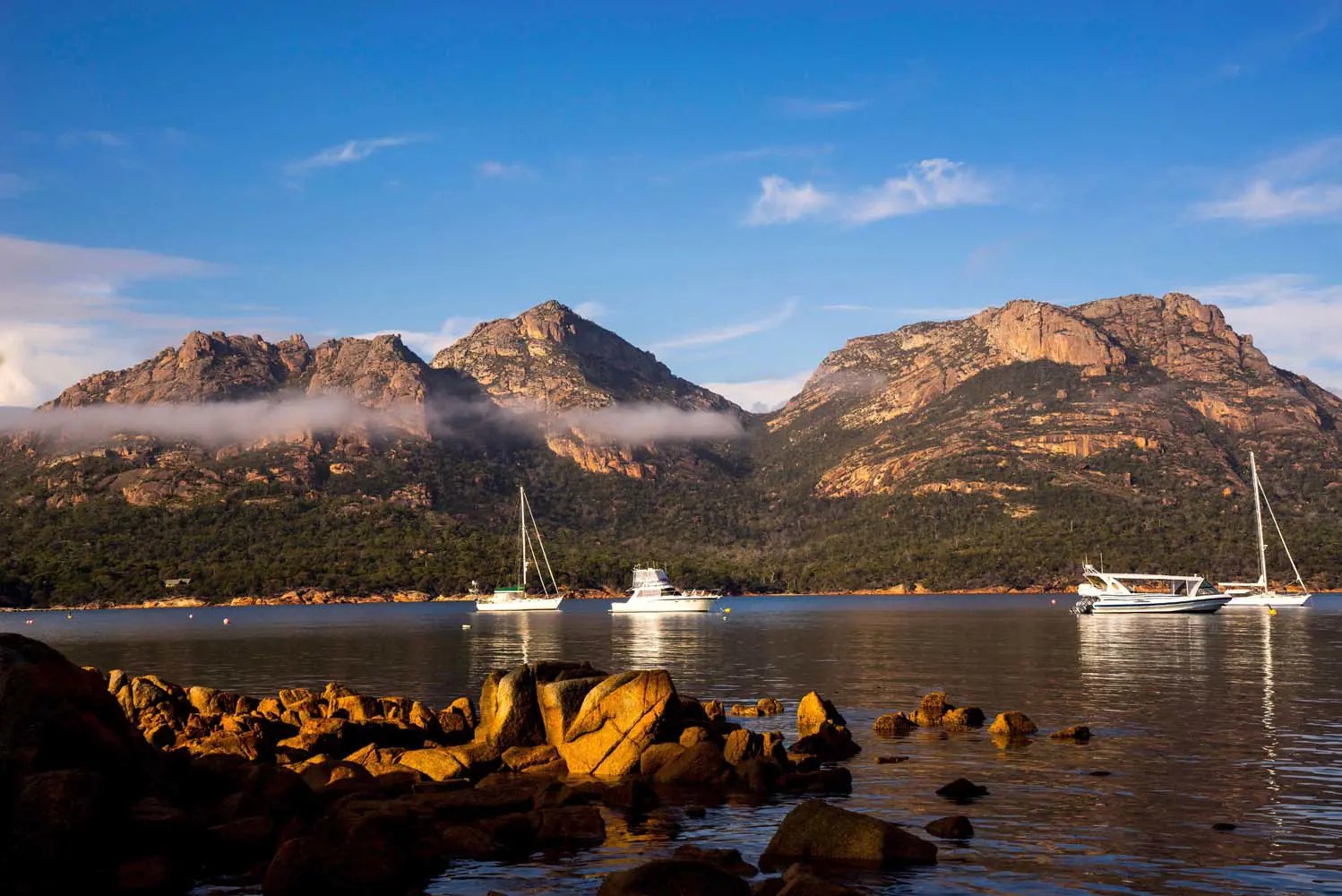 A few boats are moored on the water in a small bay, with a sharply rising mountain range towering in the background. There are a few wisps of cloud.