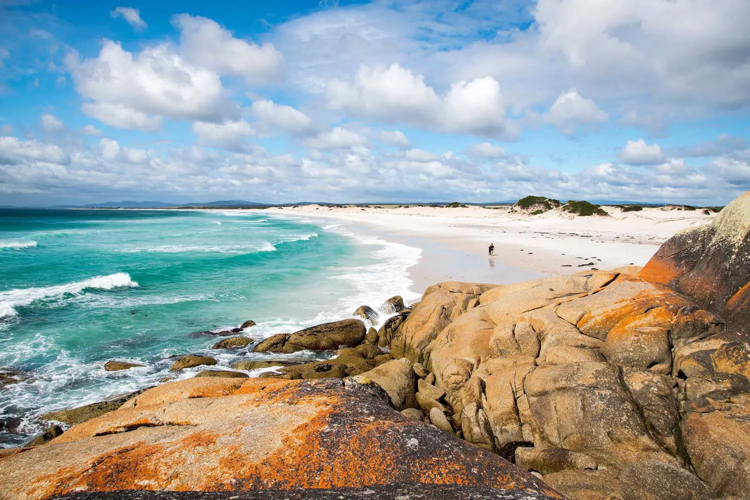 Teal waves crash onto a white expanse of beach, framed by blue sky and fluffy clouds overhead, and orange-stained sandy rocks on the shore.