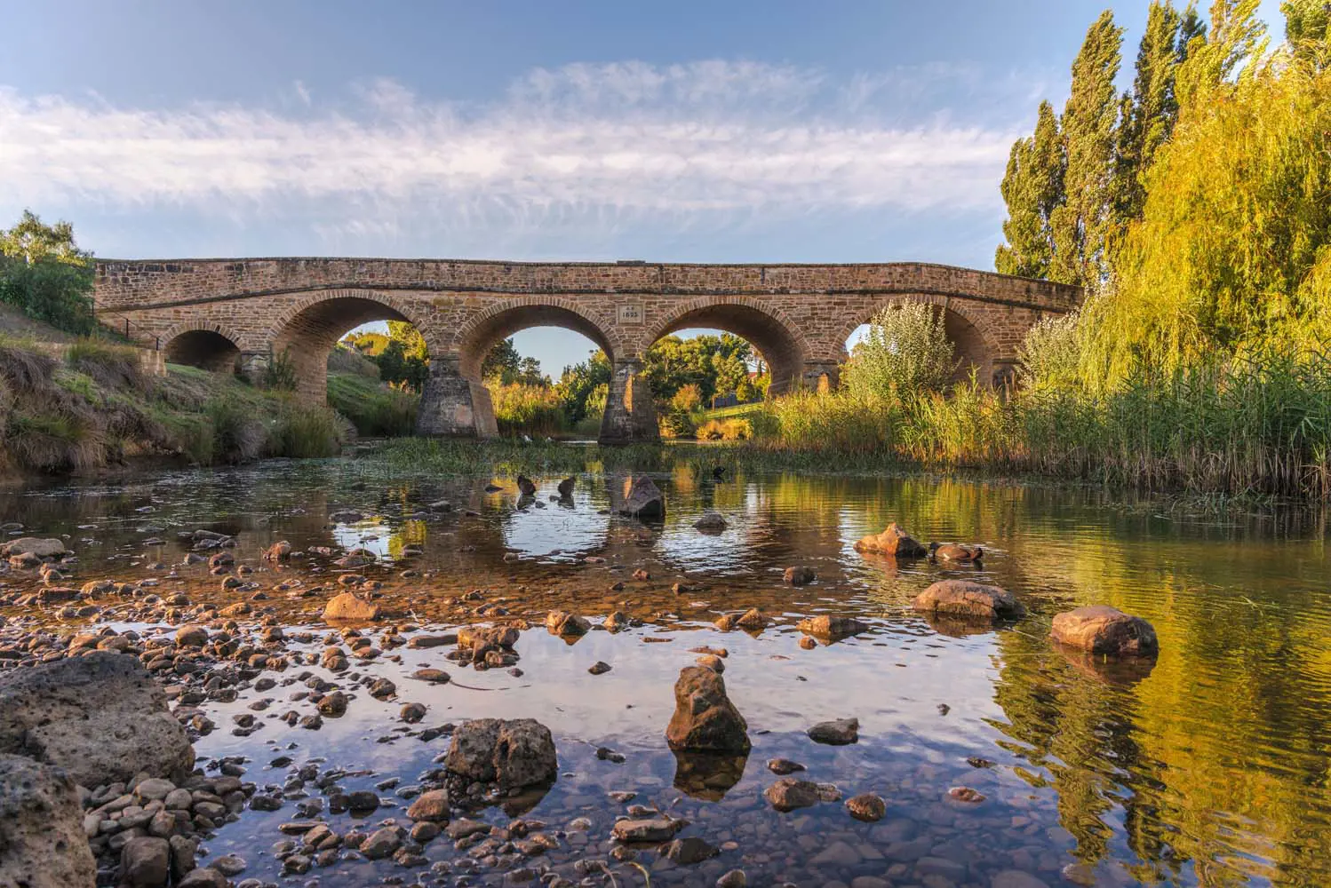 A historic stone bridge arches over a calm river. Orange-tinged trees line the banks and the very shallow water is full of pebbles and rocks.