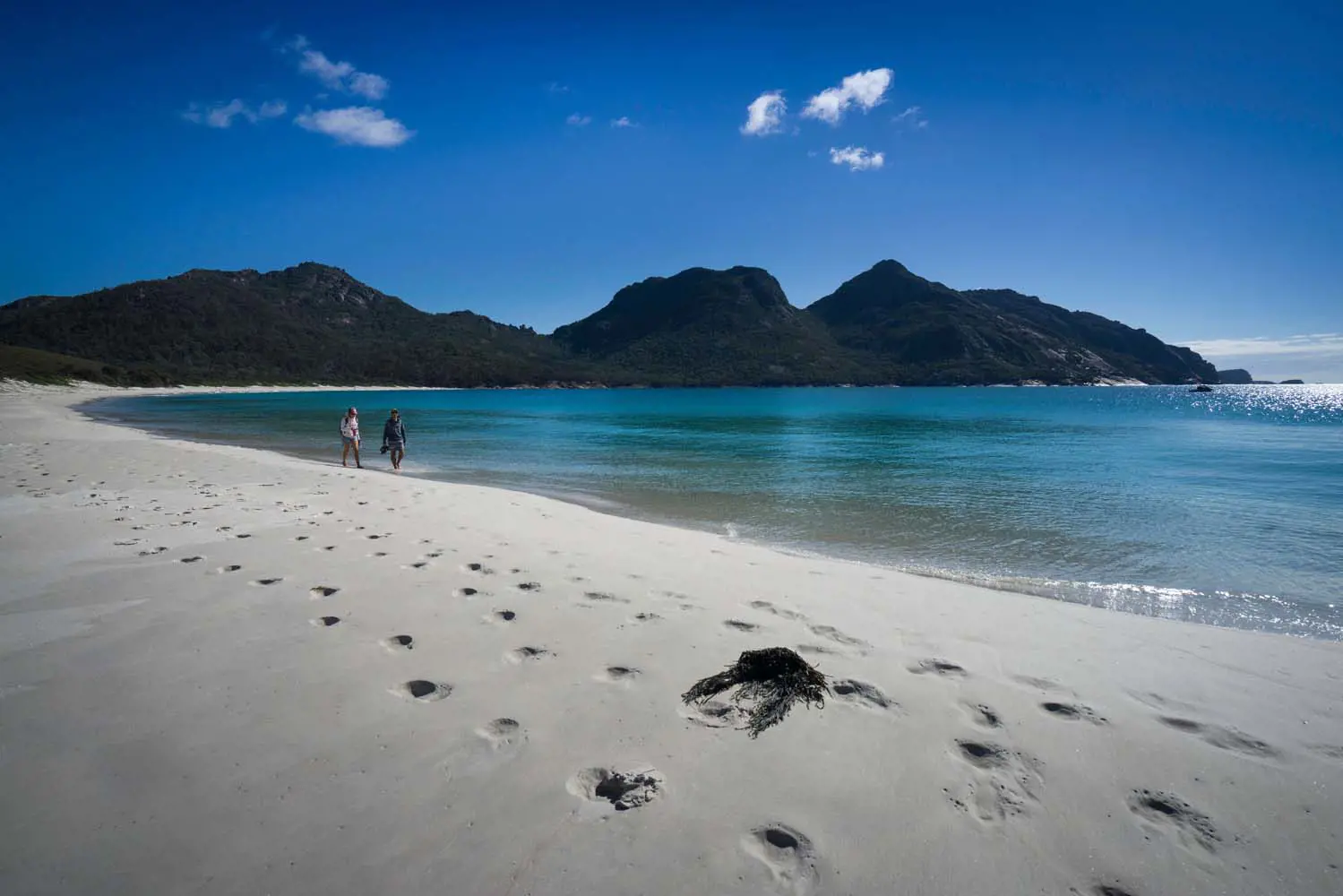 A trail of footprints through otherwise pristine white sand of a beach with clear flat ocean water and dark mountains rising in the distance.