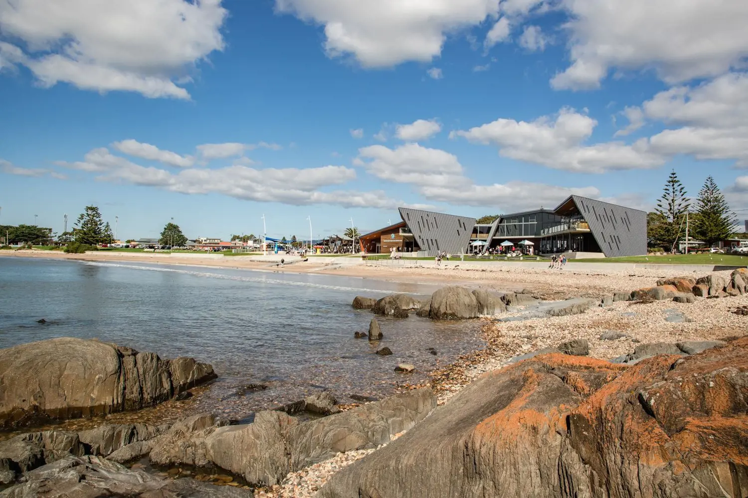 From some rocks to the side of a shallow beach, looking out to the shore where people sit on a grassed area around angular architectural buildings.