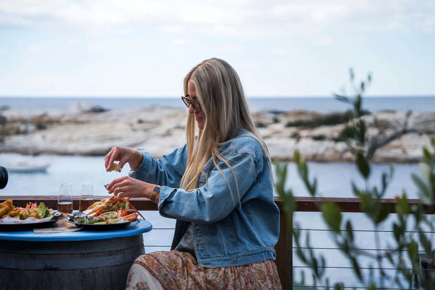 A woman in a denim jacket and sunglasses sits on an outdoor deck overlooking water, tucking into a plate full of delicious lobster and other seafood.