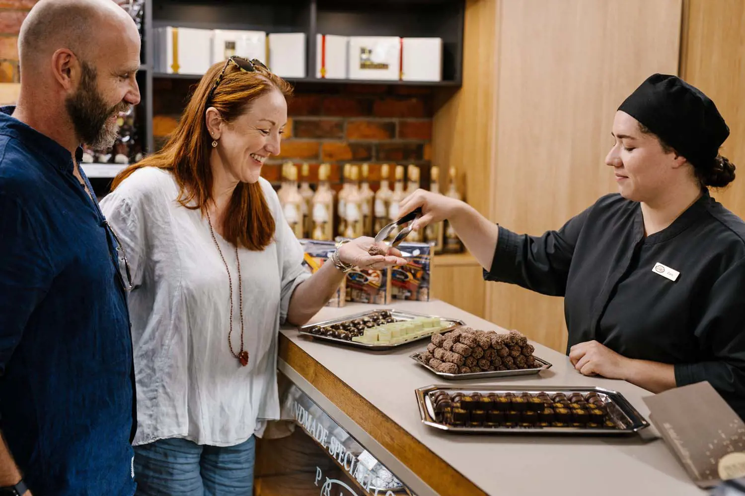 Two people stand at the counter of a chocolate shop while a staff member uses tongs to hand them a sample of chocolate to taste.